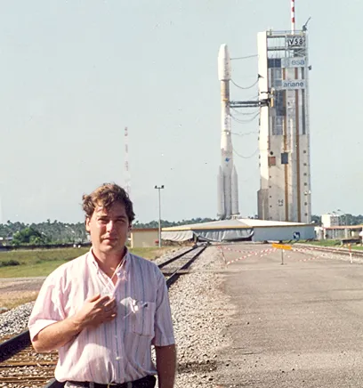 Man standing on a paved path near railway tracks with a rocket on a launch pad in the background during daytime.