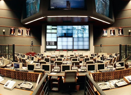 Control room with multiple computer monitors, telephones, and communication devices arranged in curved rows facing a large central video wall displaying data and images.
