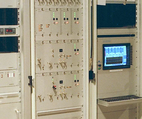A close-up of a vintage computer control panel with several rows of switches, indicator lights, and knobs, next to a monitor displaying a graph and a keyboard below it.