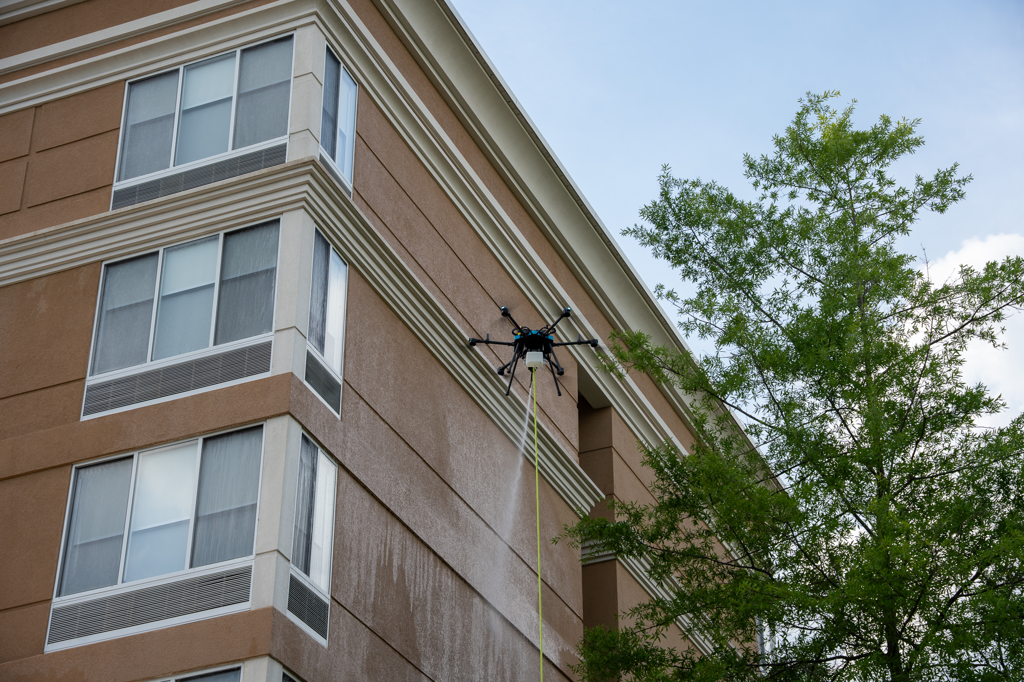 Cleaning drone soft-washing the upper corner of a multi-story hotel facade with tether line visible