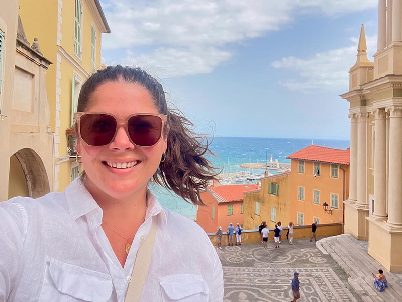 Photo of Heather. There is a street with buildings, people walking, and an ocean in the background.