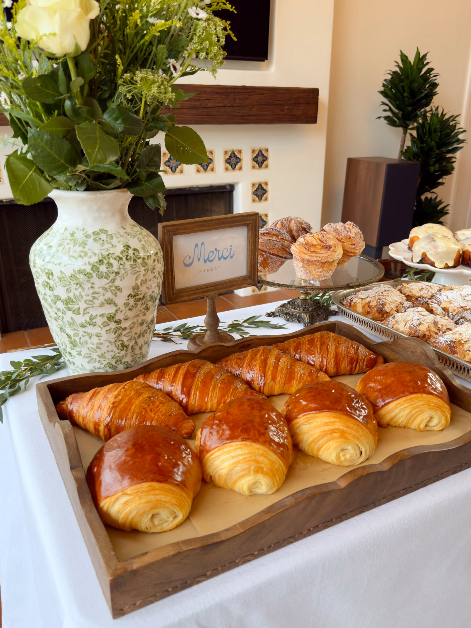 The Merci styled table: a wooden tray of croissants and pains au chocolat, a framed Merci card, cruffins on a footed stand and a vase of wildflowers on white linen