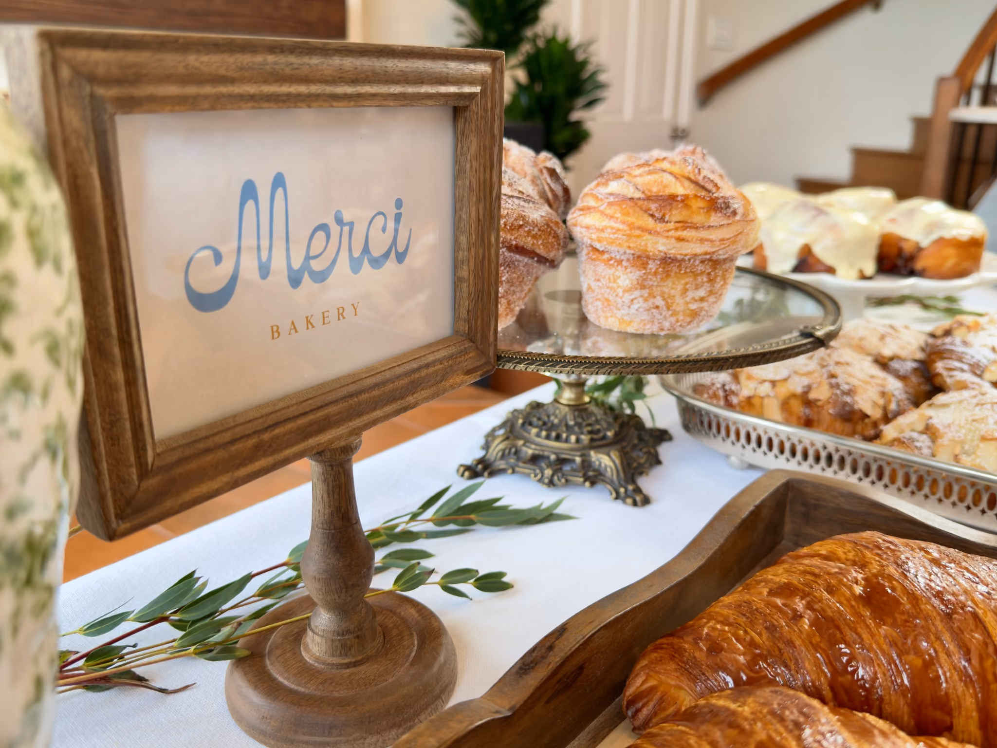 A framed Merci Bakery card beside a cruffin on a footed glass stand, with eucalyptus on the linen
