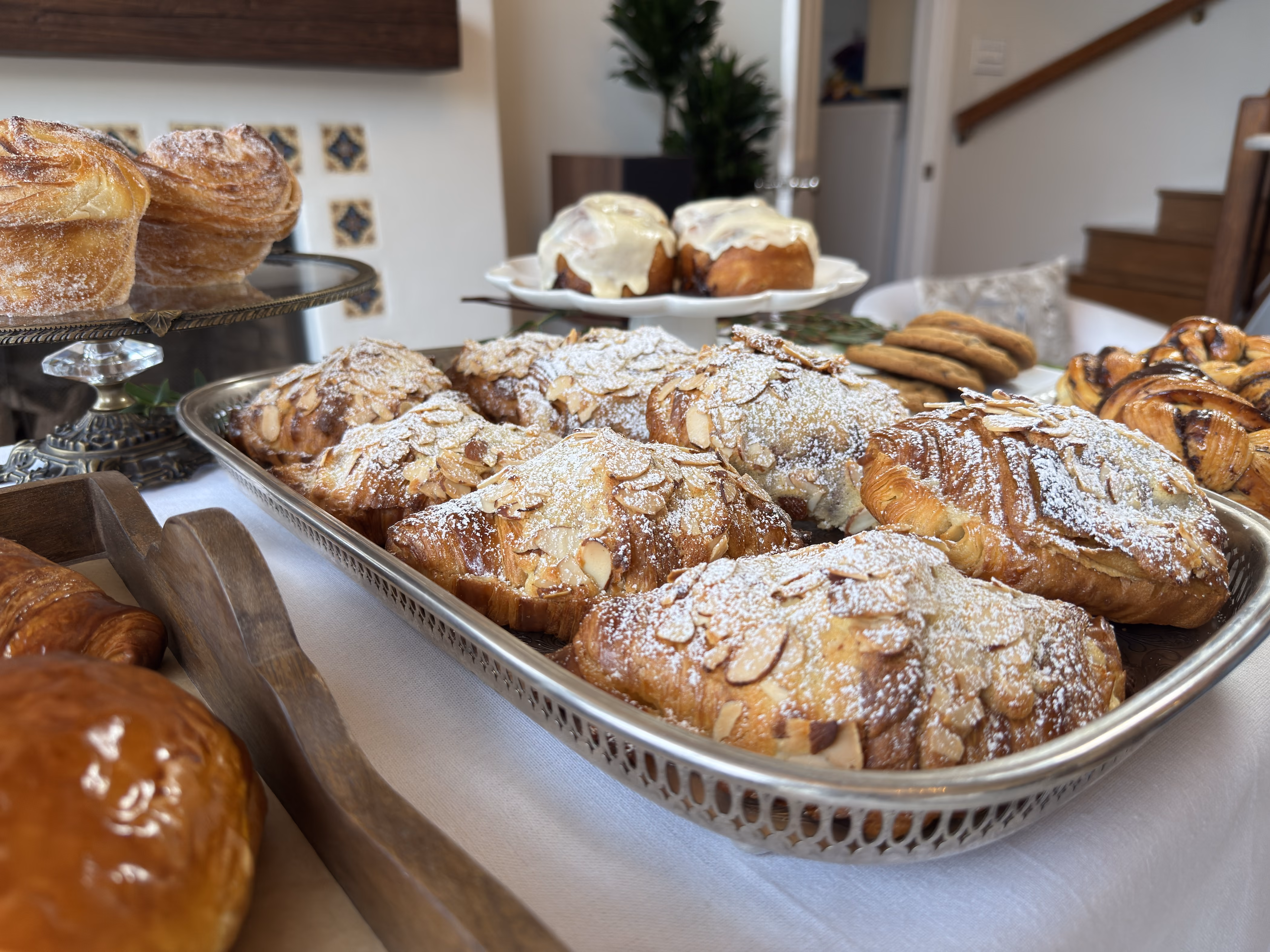 Almond croissants dusted with sugar on a silver platter, cinnamon rolls and cookies behind