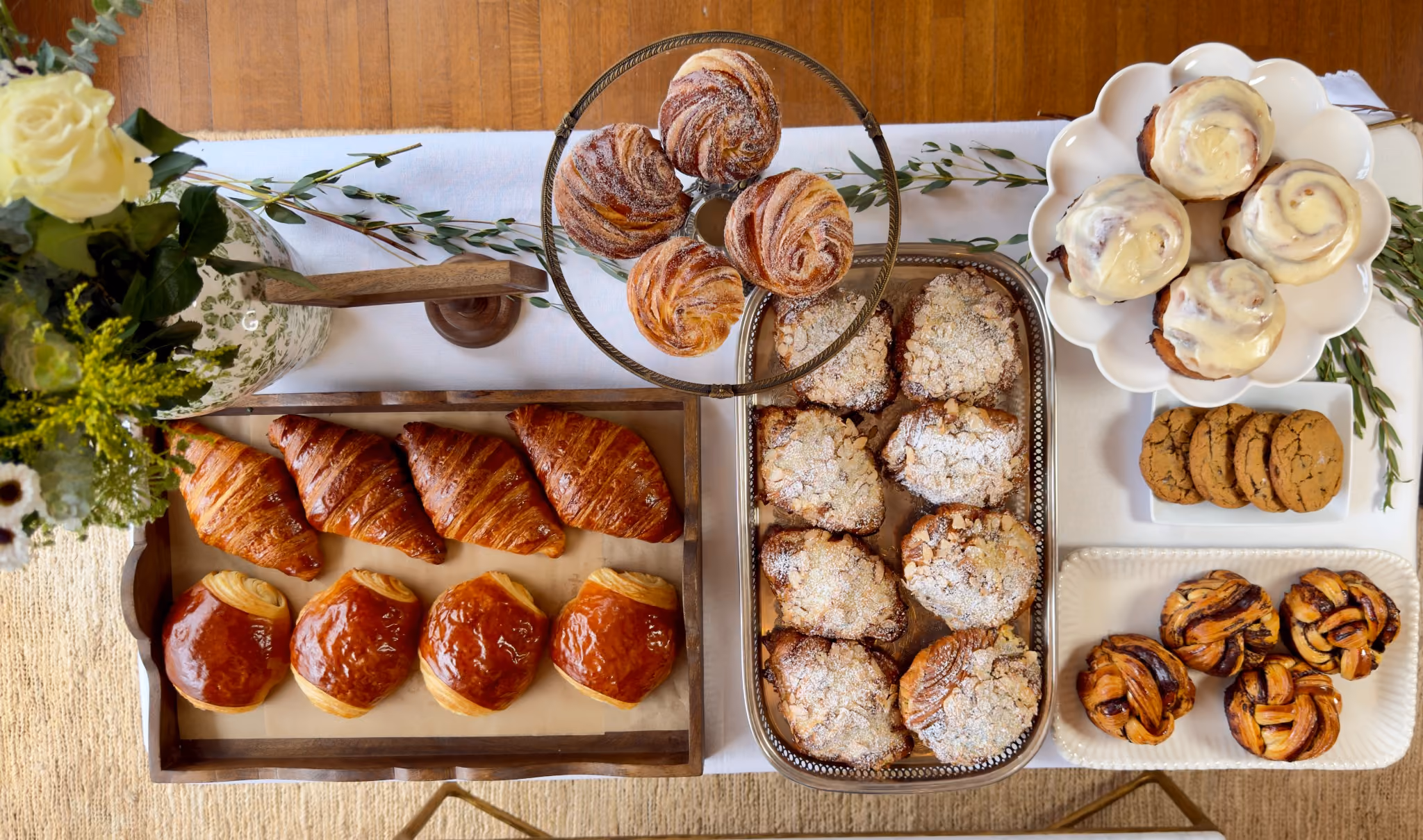 Overhead view of a Merci Bakery styled table: cinnamon rolls in a wooden crate, cruffins on a footed stand, croissants and a pot of jam on white linen