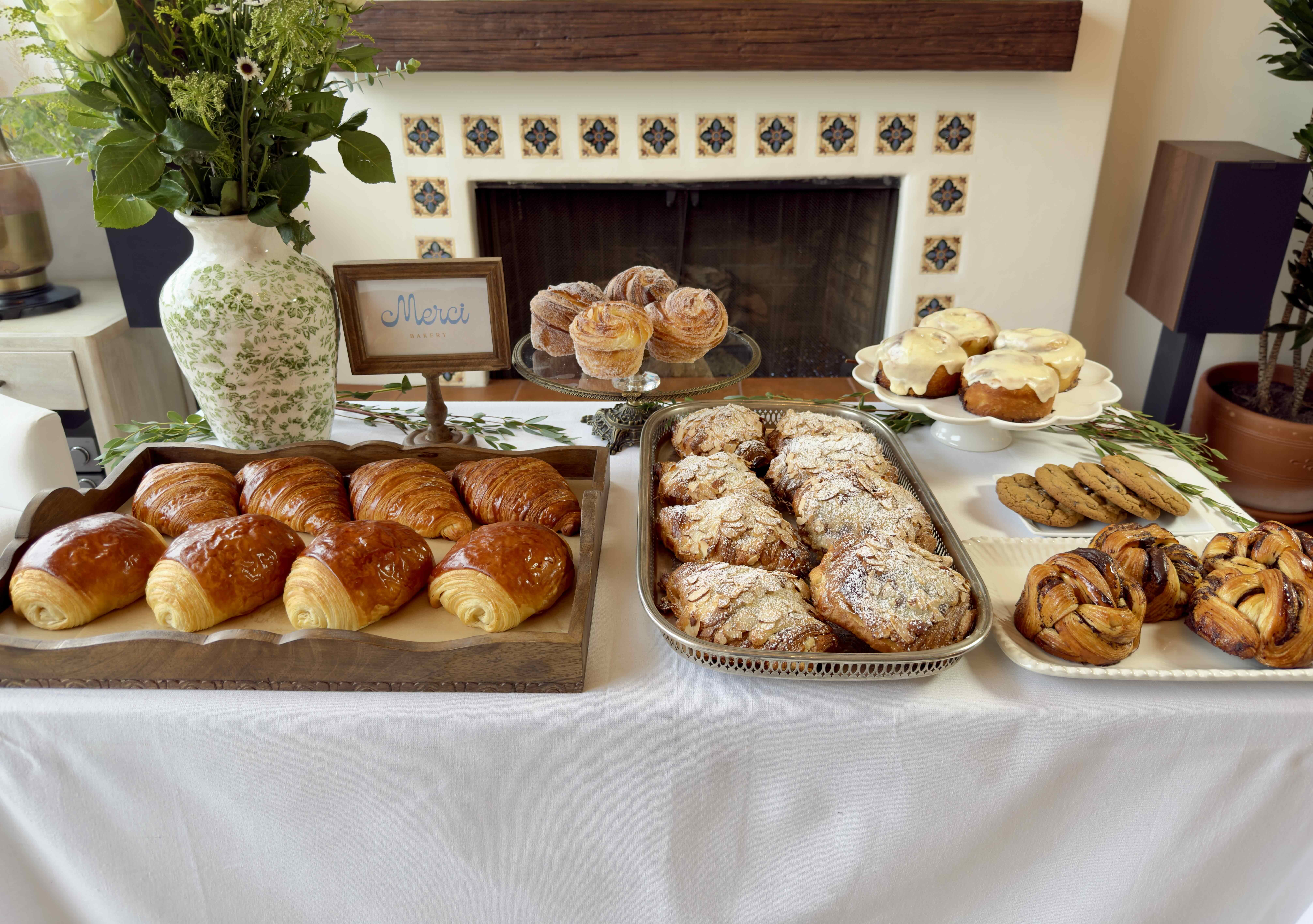 A Merci Bakery styled table dressed in white linen: trays of croissants, a silver platter of almond croissants, cinnamon rolls on a stand and flowers in a stone vase
