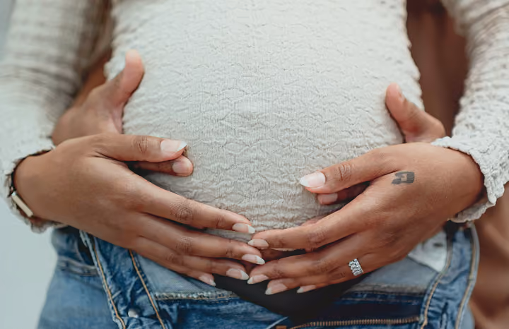 Close-up of four hands gently cradling a pregnant belly dressed in a light textured top.
