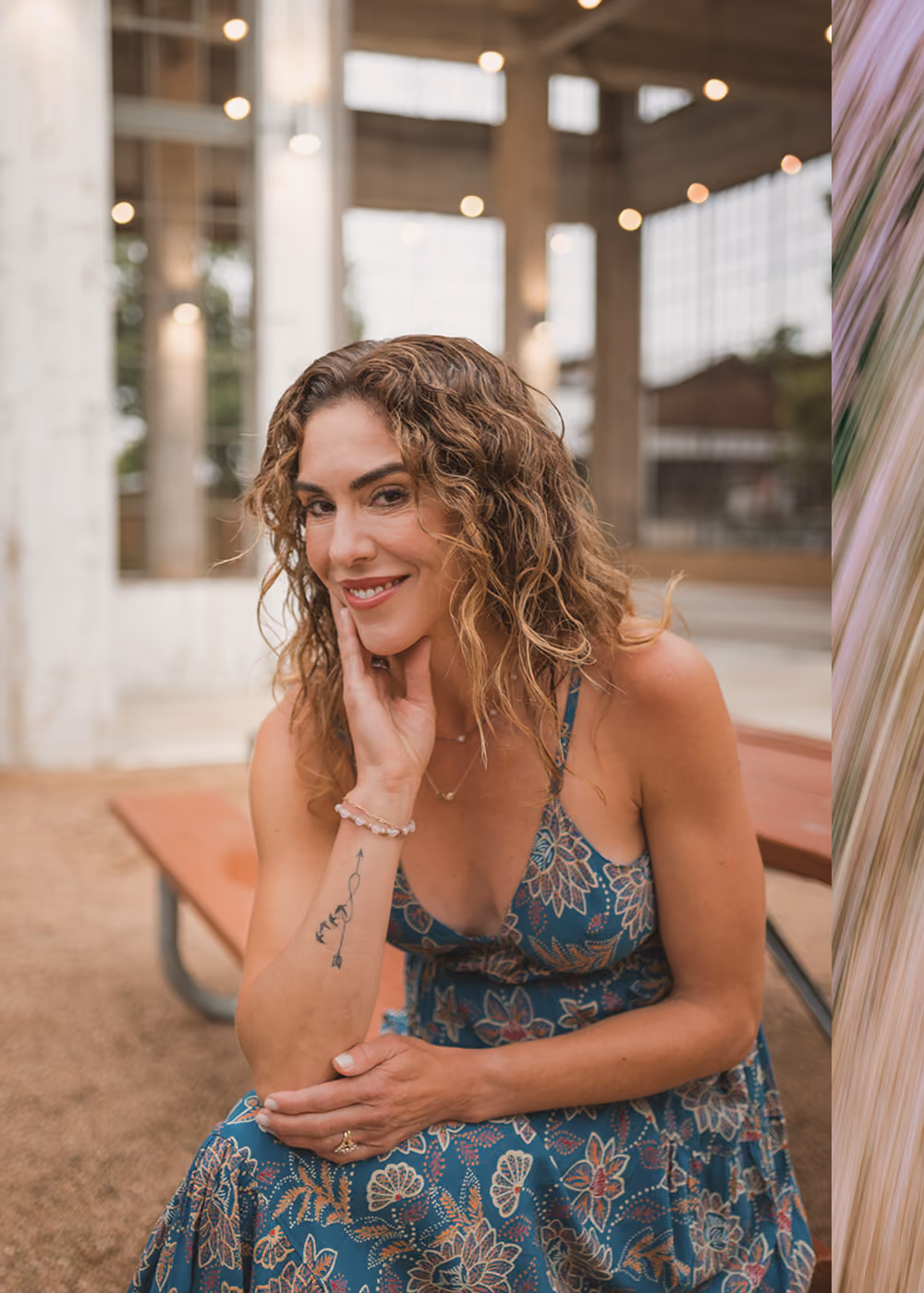 Kelly Hyde in a floral dress sitting and smiling with hand on her face and tattoo on her arm.