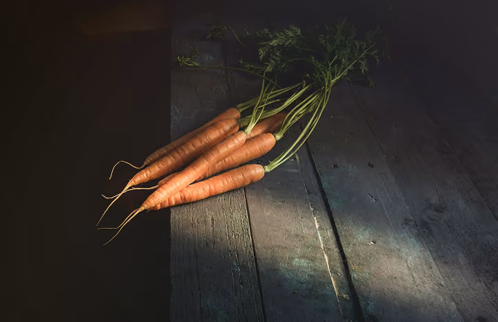 Four fresh carrots with green tops lying on a rustic wooden surface with shadow lighting.