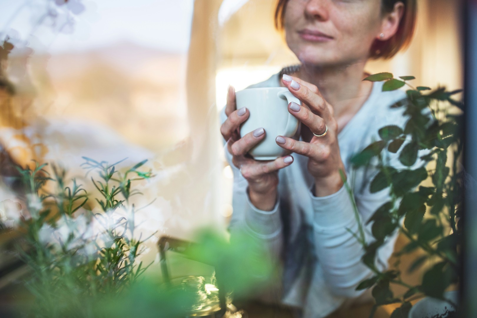 Woman holding a white mug, sitting indoors behind green plants, looking out a window thoughtfully.