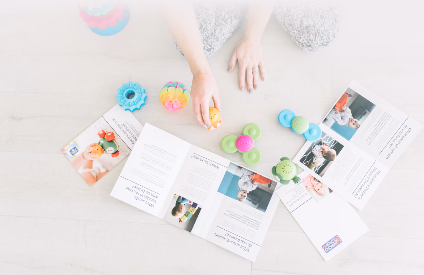 Hands sorting through colourful baby toys and product brochures spread across a white table