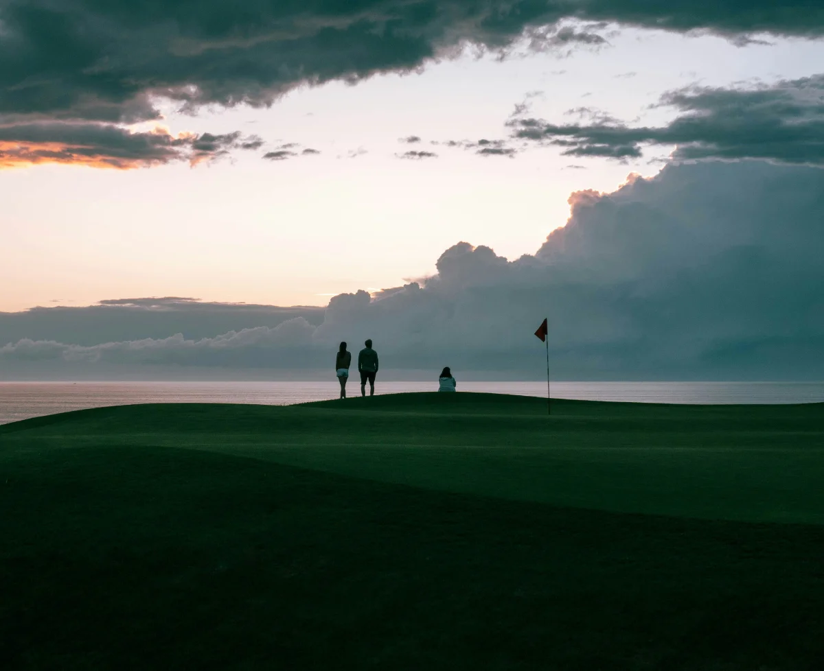 Three silhouetted figures on a cliffside green at sunset, ocean and storm clouds beyond.