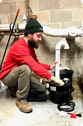 A man in a red sweatshirt and black hat working on plumbing pipes in a basement.