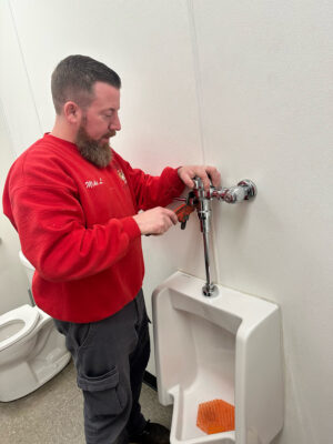 Person in red shirt using grinder tool on a grey metal pipe in a wall.