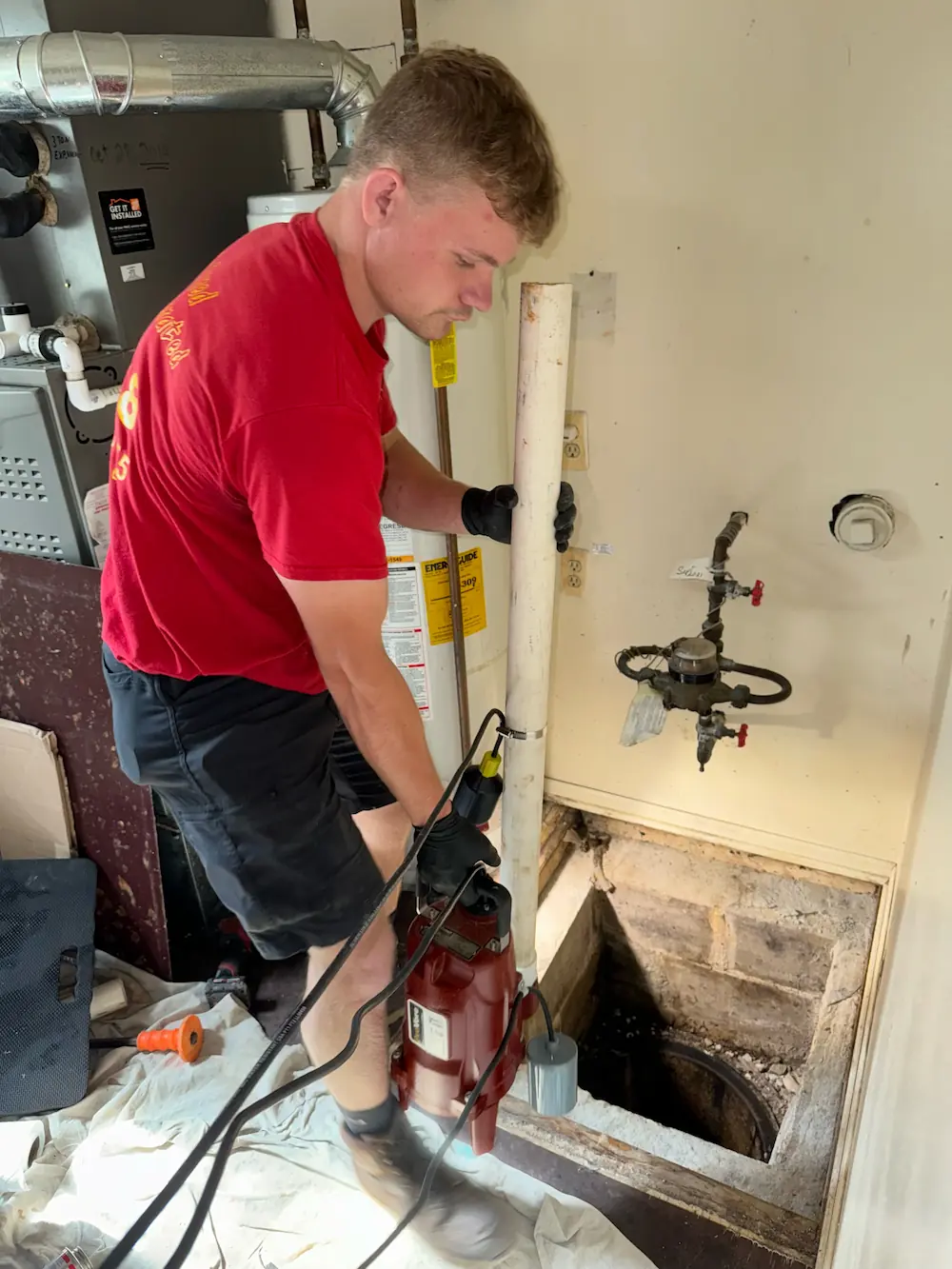 Man in a red shirt uses a level tool on pipes near two water heaters in a basement.