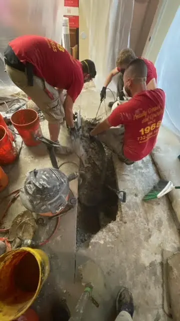 Plumber in red shirt and black cap inspecting a water heater with a flashlight in a utility room.