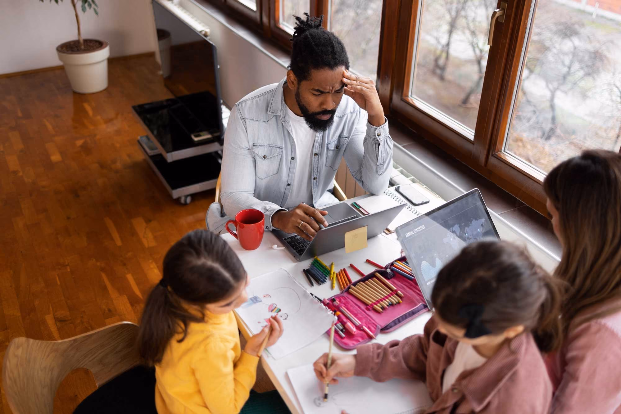 Man working on laptop at table with two children drawing and a woman nearby by a window.