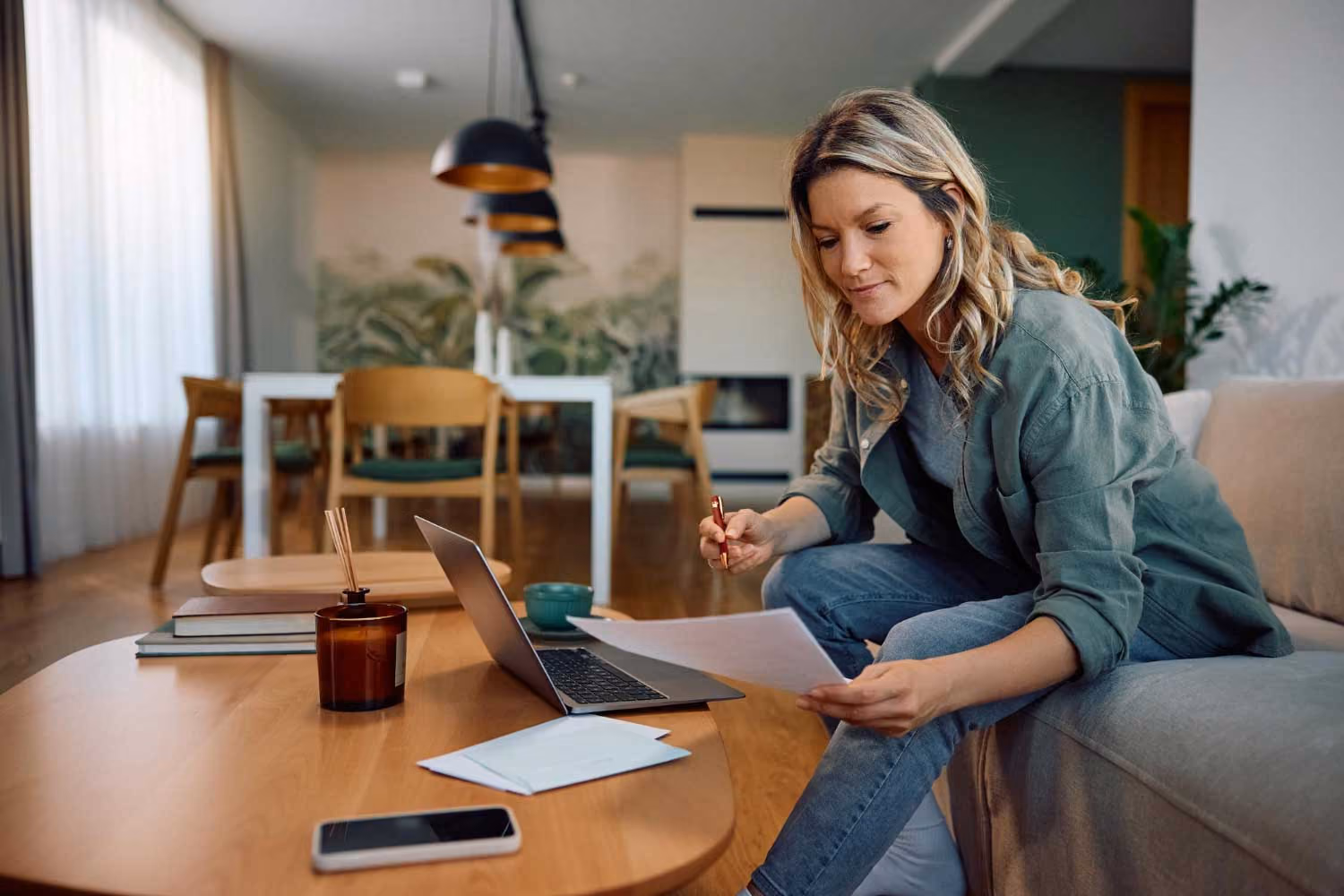 Woman sitting on a sofa reviewing papers with a laptop and phone on a coffee table.