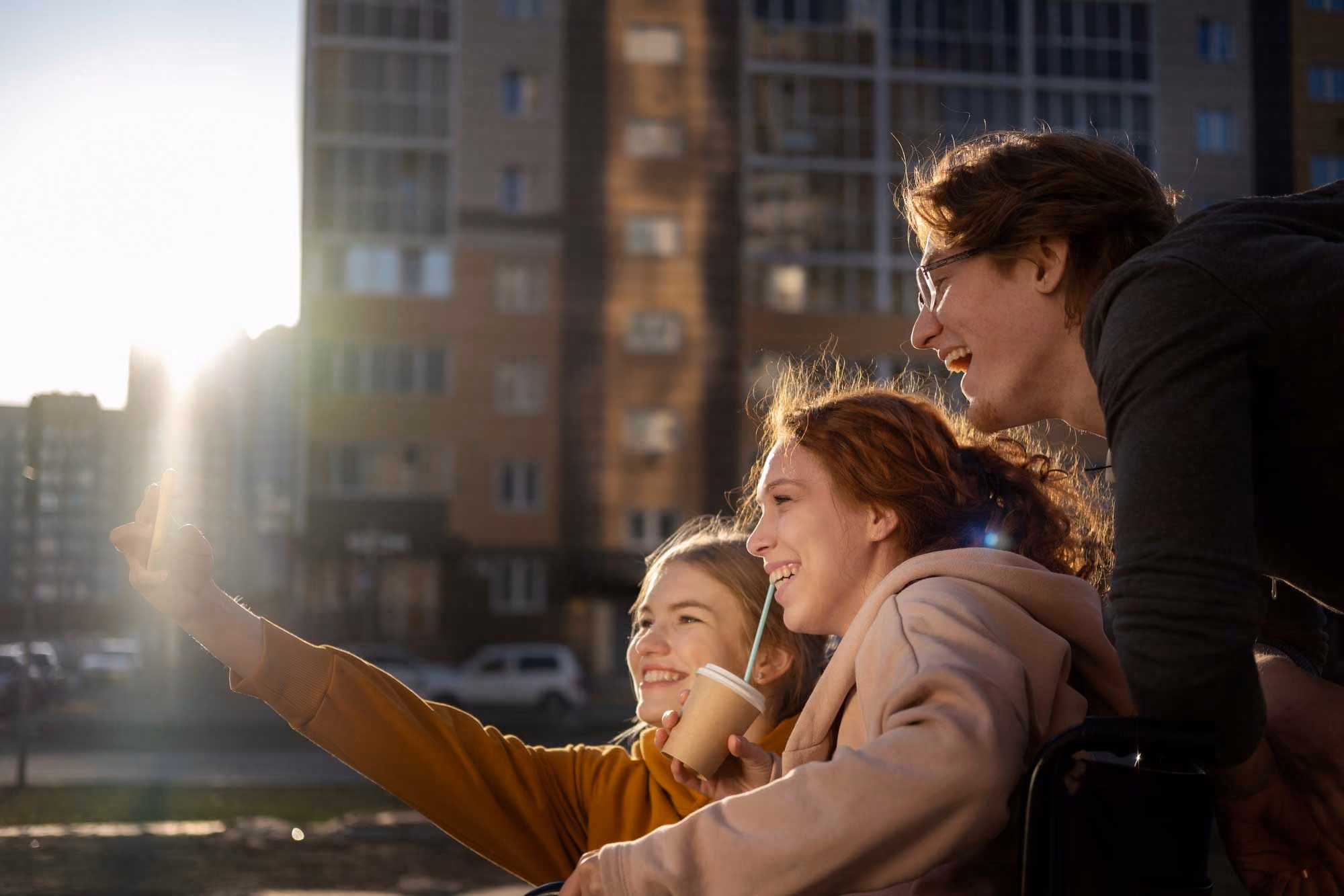 Three friends smiling and taking a selfie outdoors near buildings during sunset.