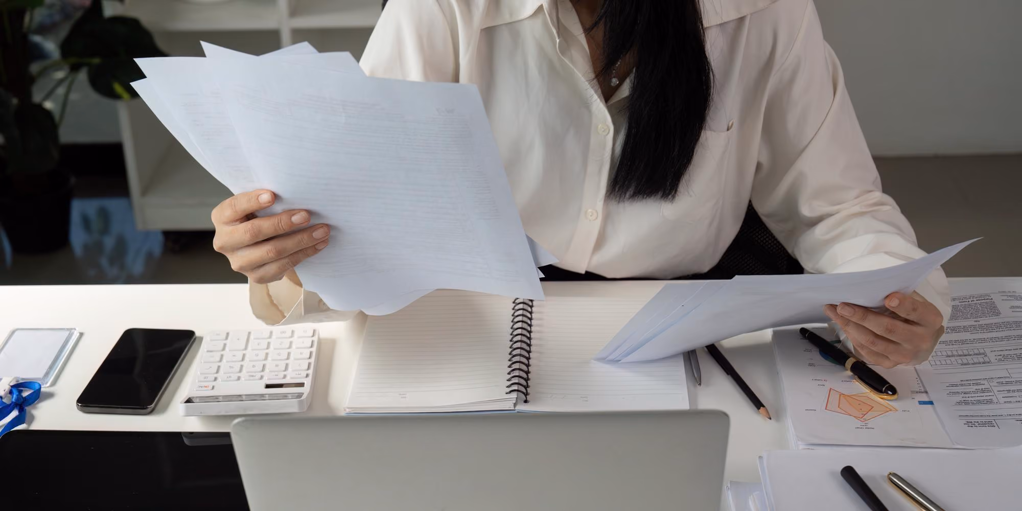 Person in white shirt sorting through papers at desk with notebook, phone, calculator, and laptop.