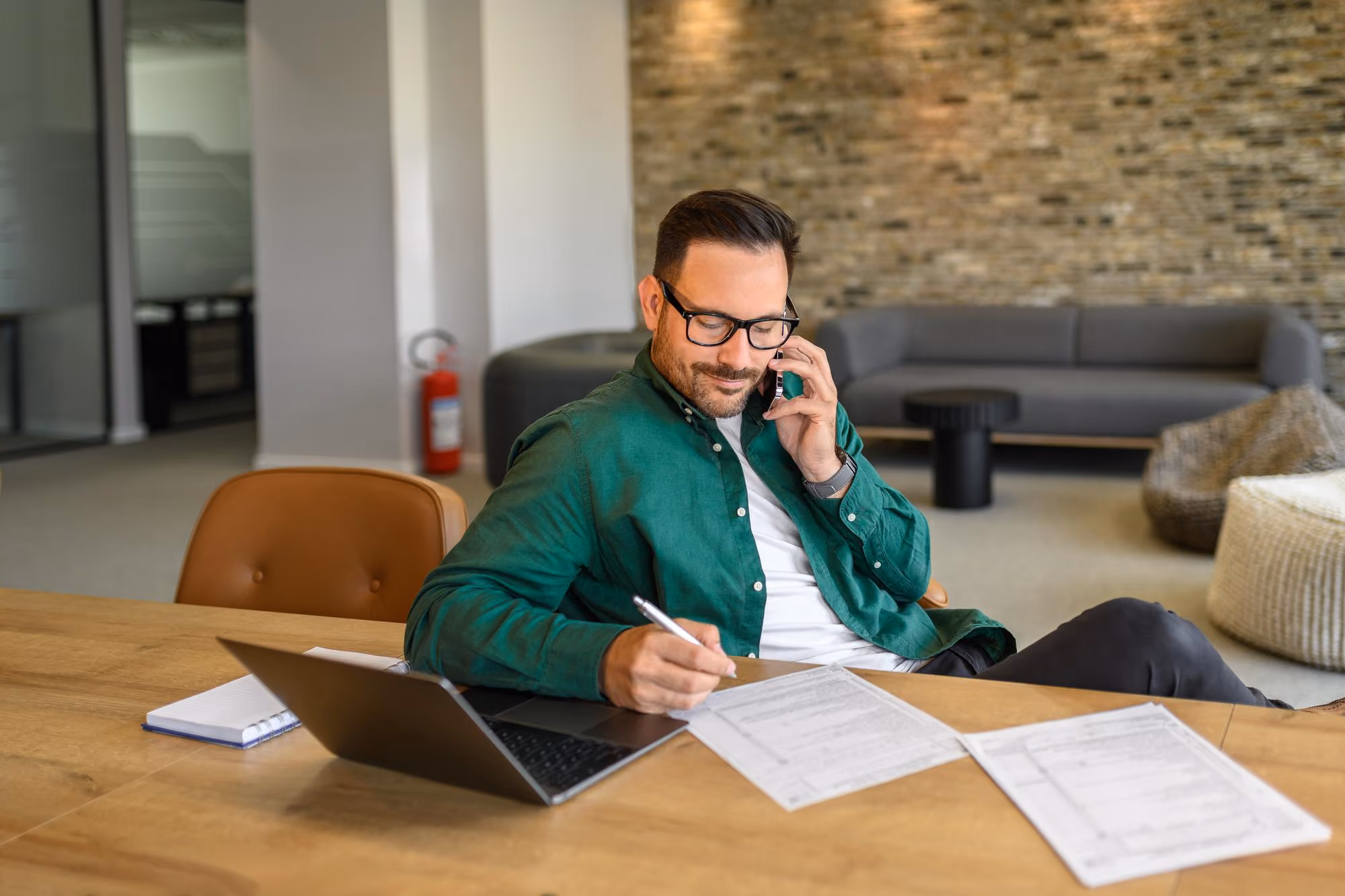 Man in green shirt sitting at desk, talking on phone while writing on paperwork with laptop open.
