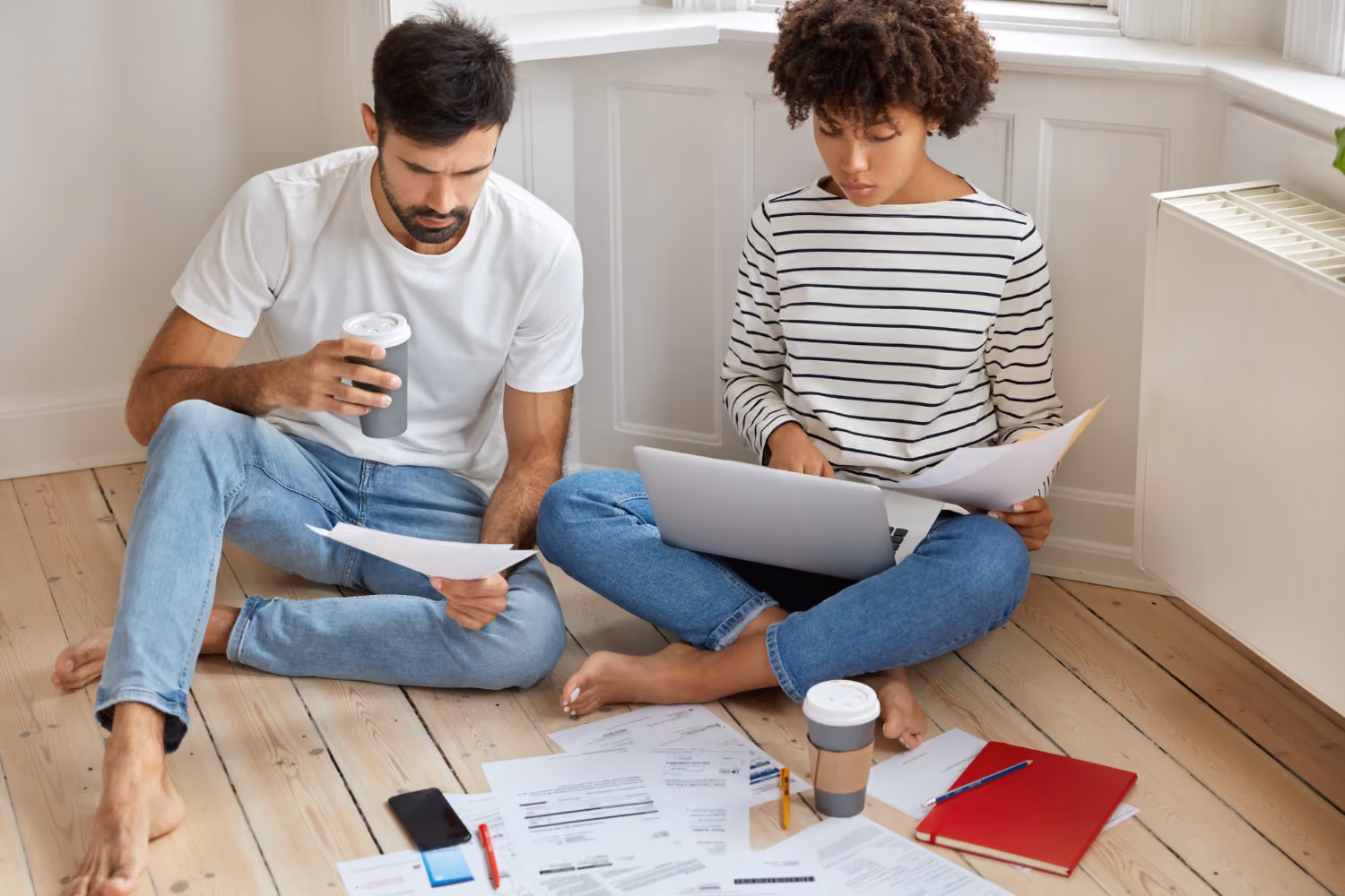 Man and woman sitting on floor surrounded by papers, reviewing tax abatement documents with laptop and coffee cups.