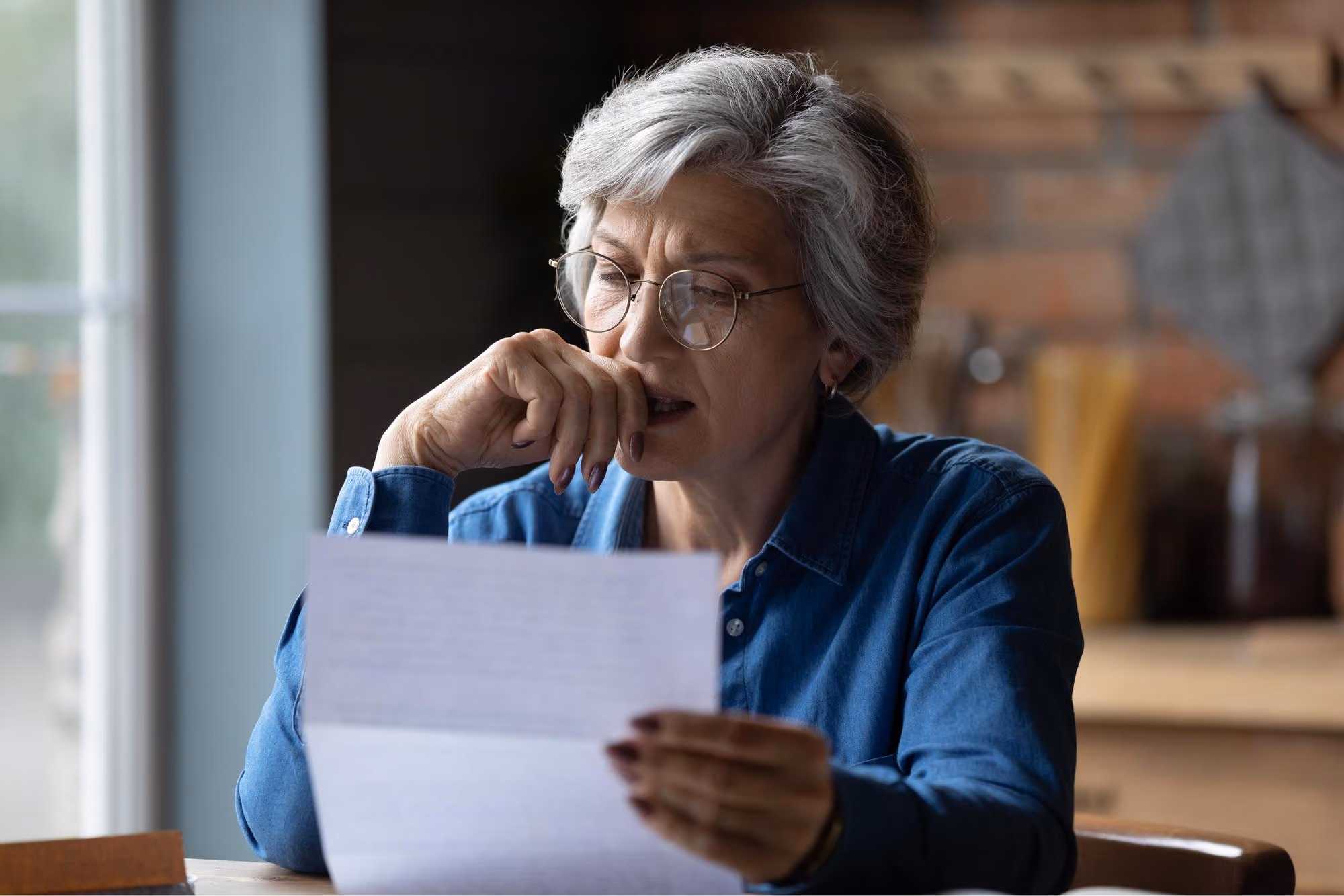 Older woman with glasses in blue shirt reading a letter with a thoughtful expression indoors.