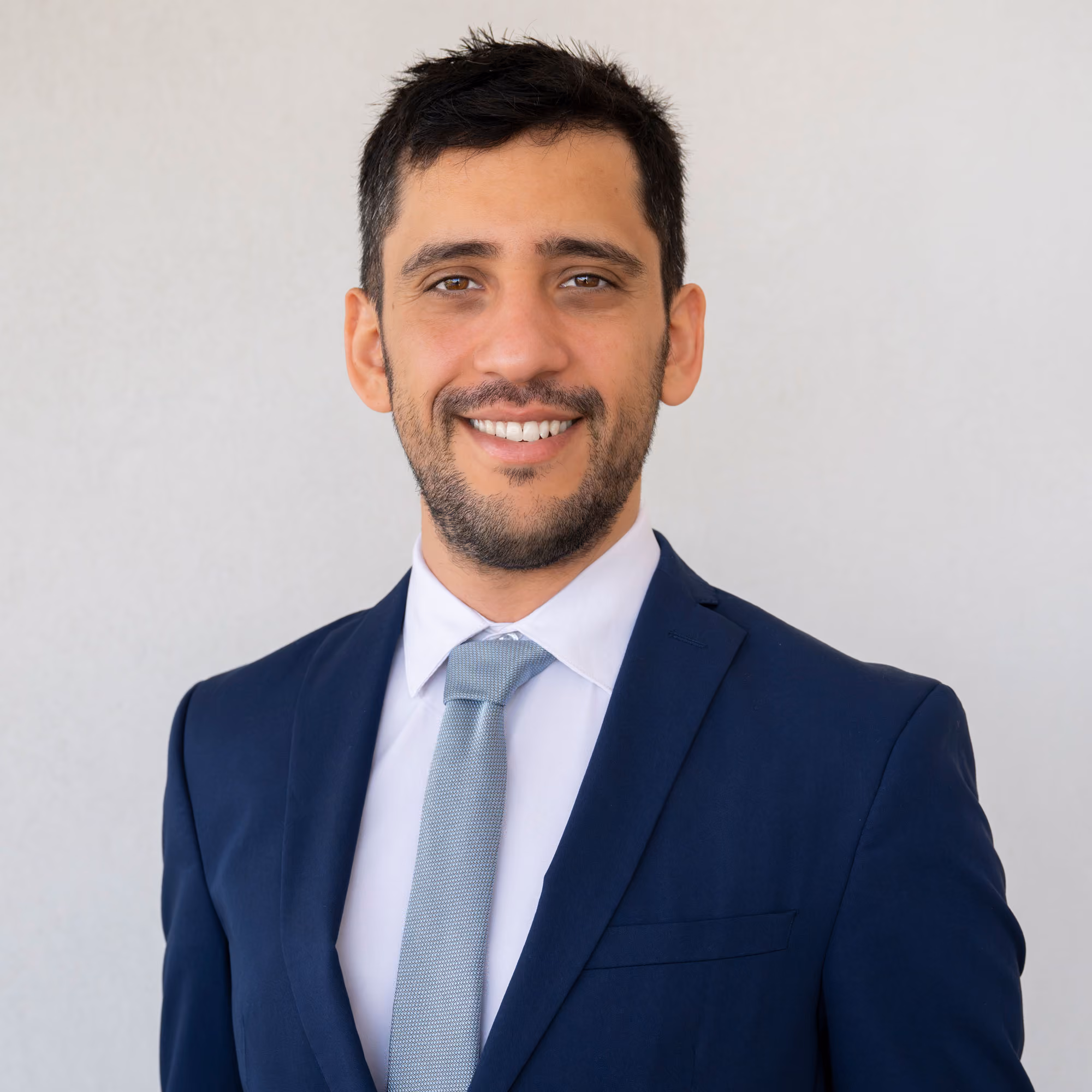 A smiling Ori Efroni, founder of NYC TaxAbate Advisors with dark hair wearing a navy suit, white shirt, and light gray tie against a plain background.