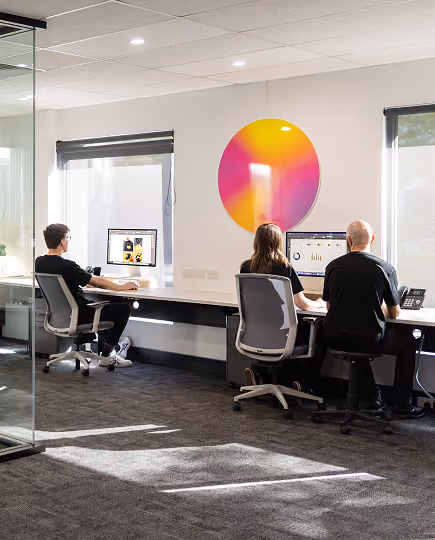 Three people working on desktop computers in a brightly lit modern office with large windows and a colorful circular wall decoration.