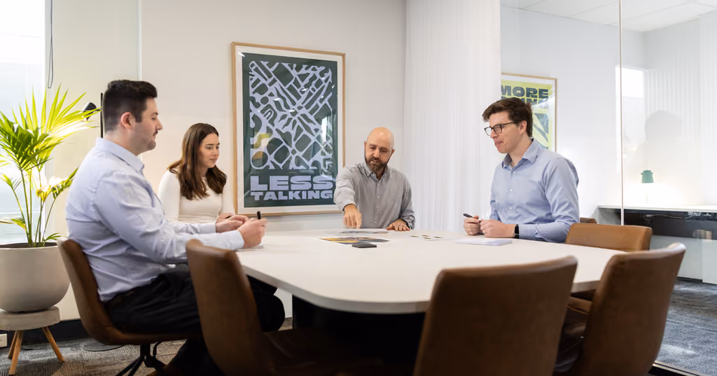 Four people in a meeting around a table with posters reading 'LESS TALKING' and 'MORE ACTION' behind them.