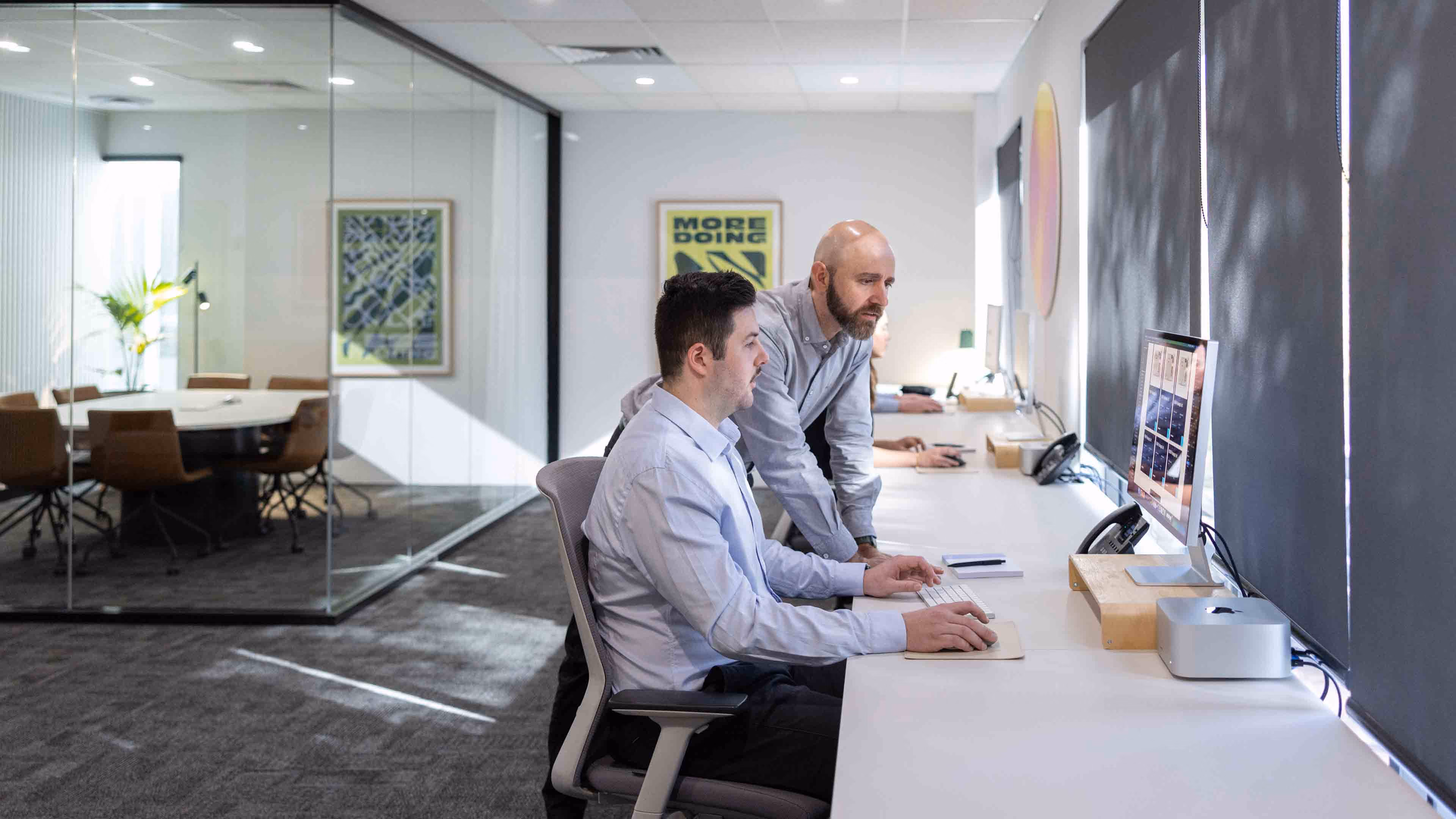 Two men collaborating at a computer workstation in a modern office with glass walls and blinds.