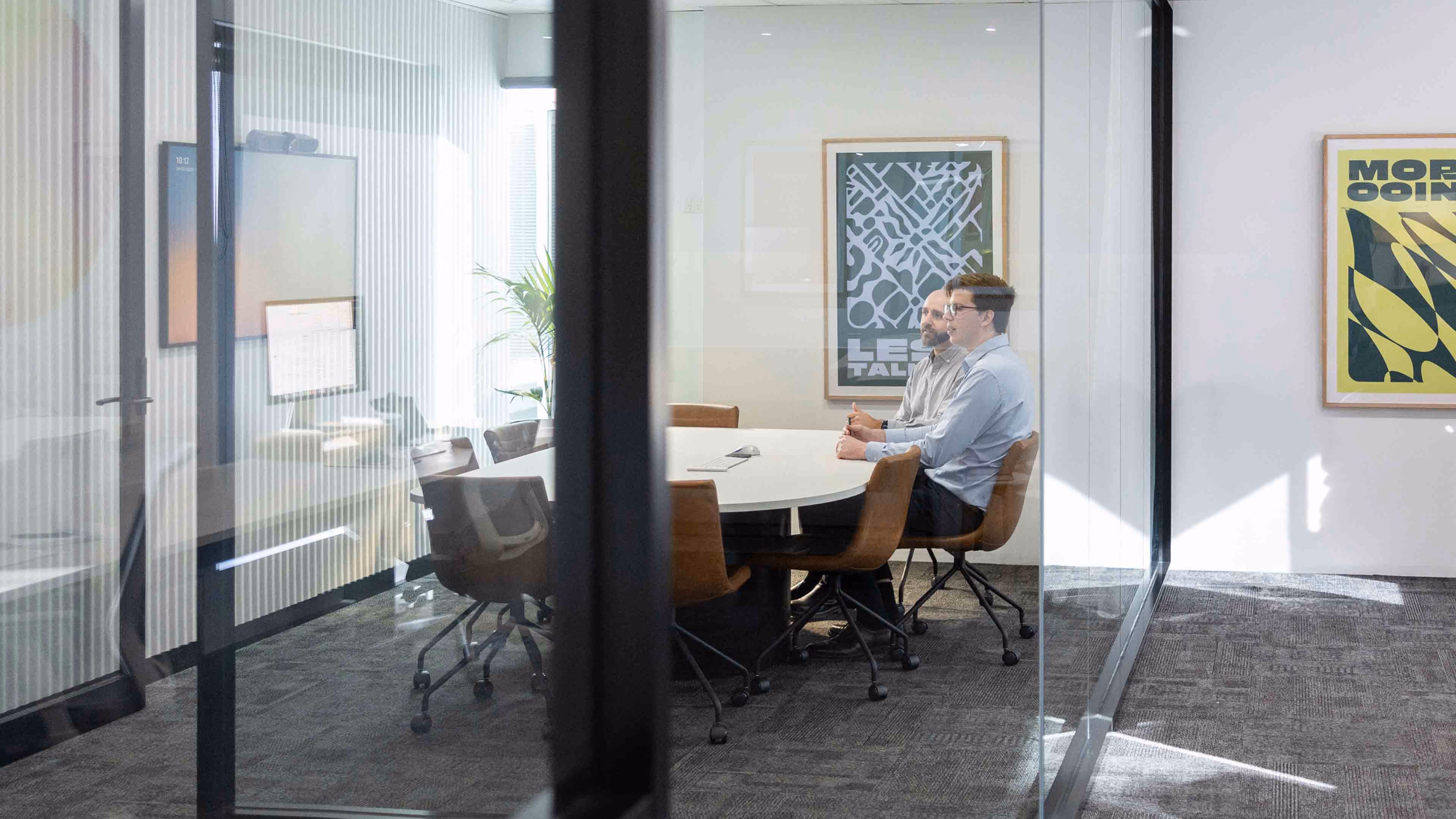 Two men sitting and talking at a round table in a glass-walled office meeting room.