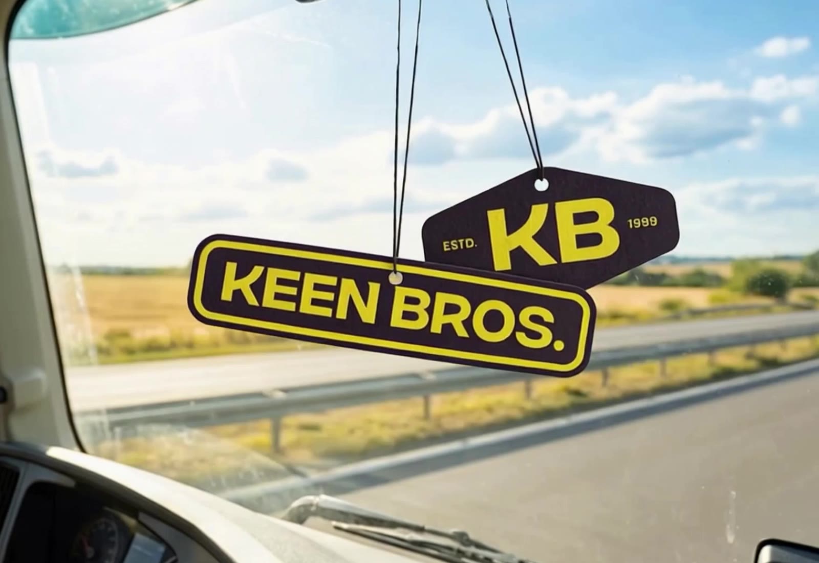 Two hanging car air fresheners read KB and KEEN BROS in yellow on black with roadside view.