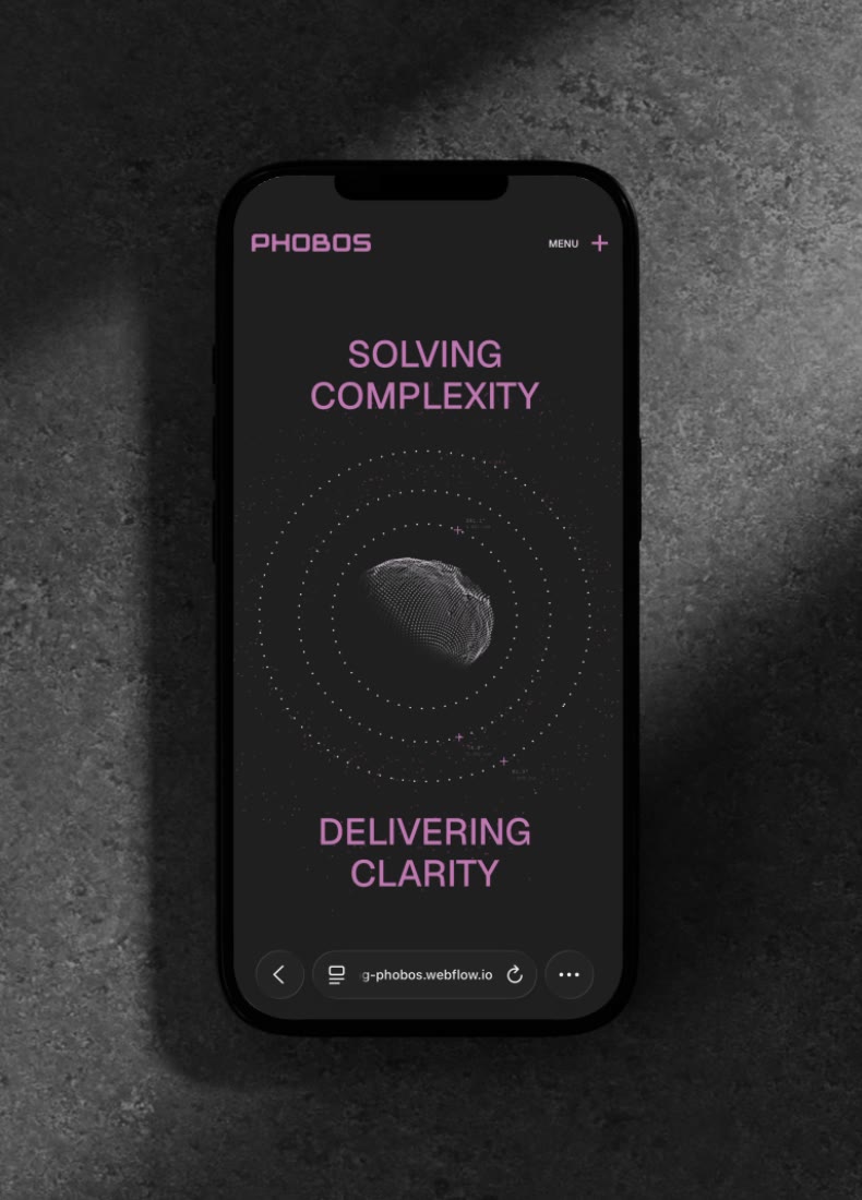 Black smartphone on dark surface showing PHOBOS site with text 'Solving Complexity Delivering Clarity' and a dotted globe.