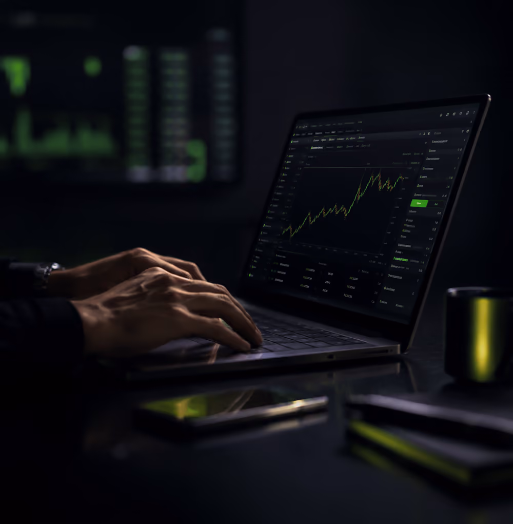 Hands typing on a laptop showing stock market charts and data on screen in a dark room.