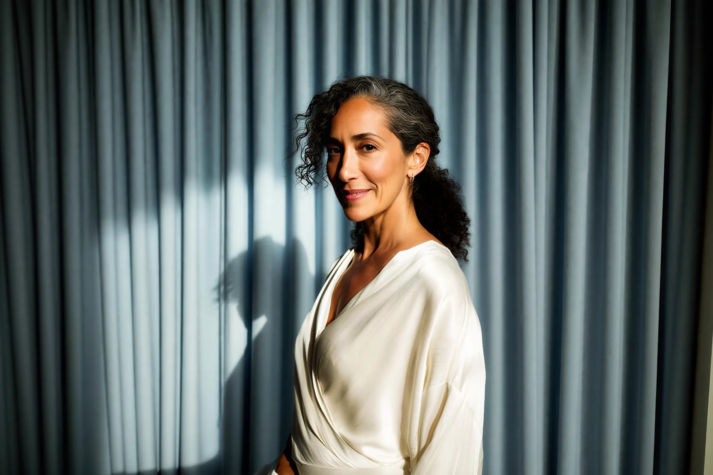 Smiling woman with curly salt-and-pepper hair wearing a white satin blouse standing in soft light against gray curtains.