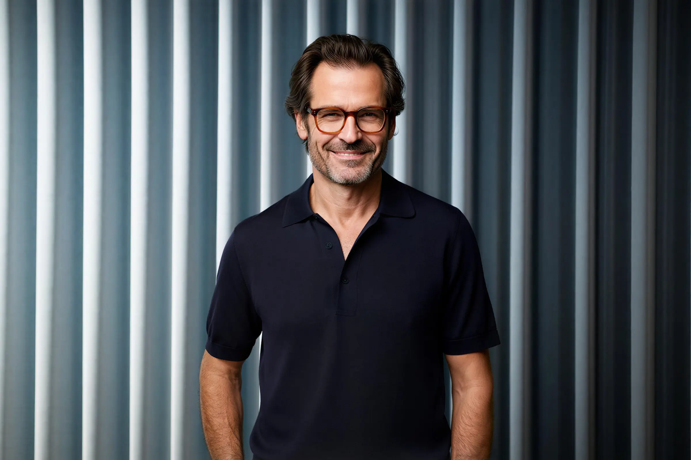 Smiling middle-aged man with glasses wearing a dark polo shirt in front of a textured gray curtain background.