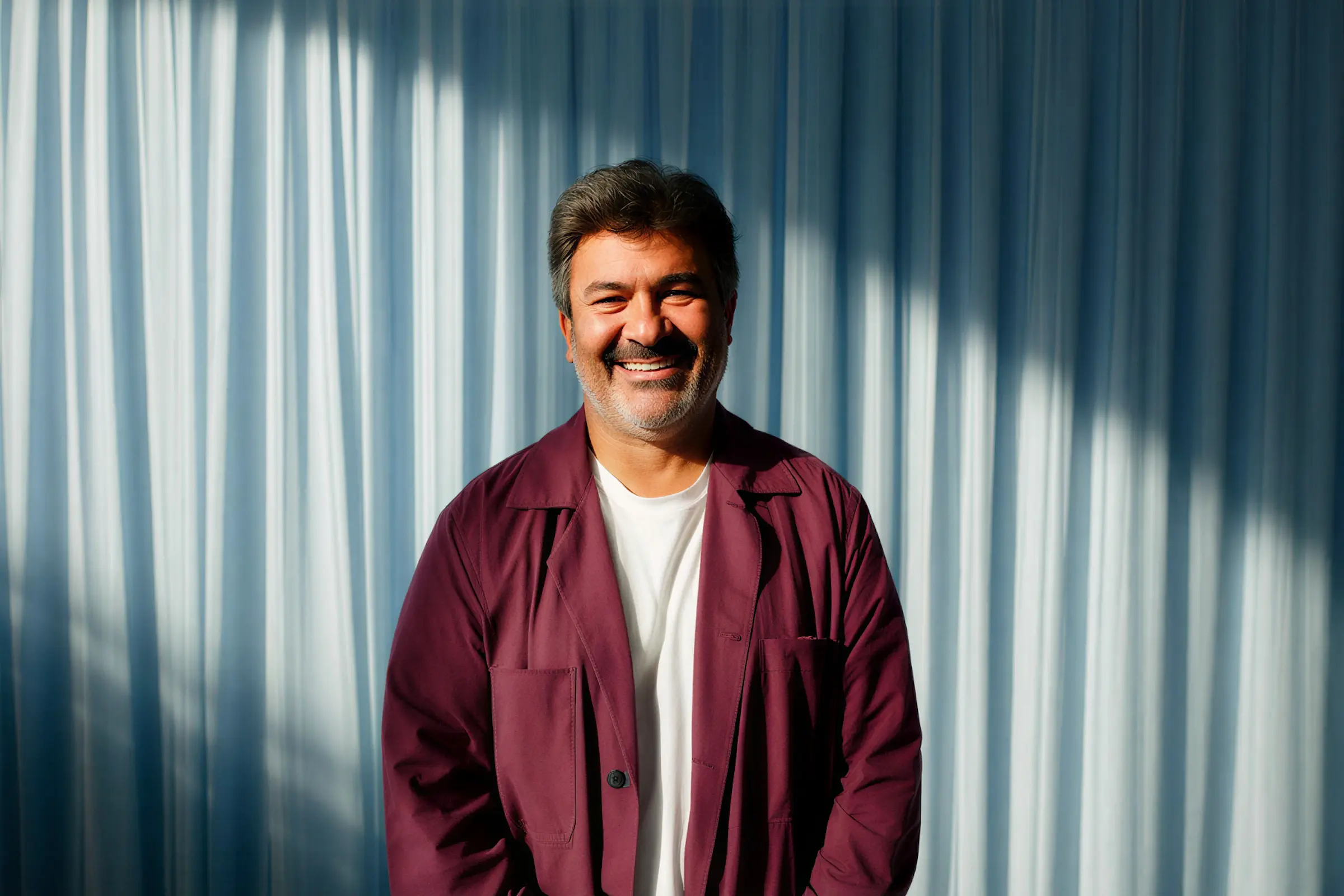 Smiling middle-aged man with salt-and-pepper hair and beard wearing a maroon jacket and white shirt in front of light blue curtains.
