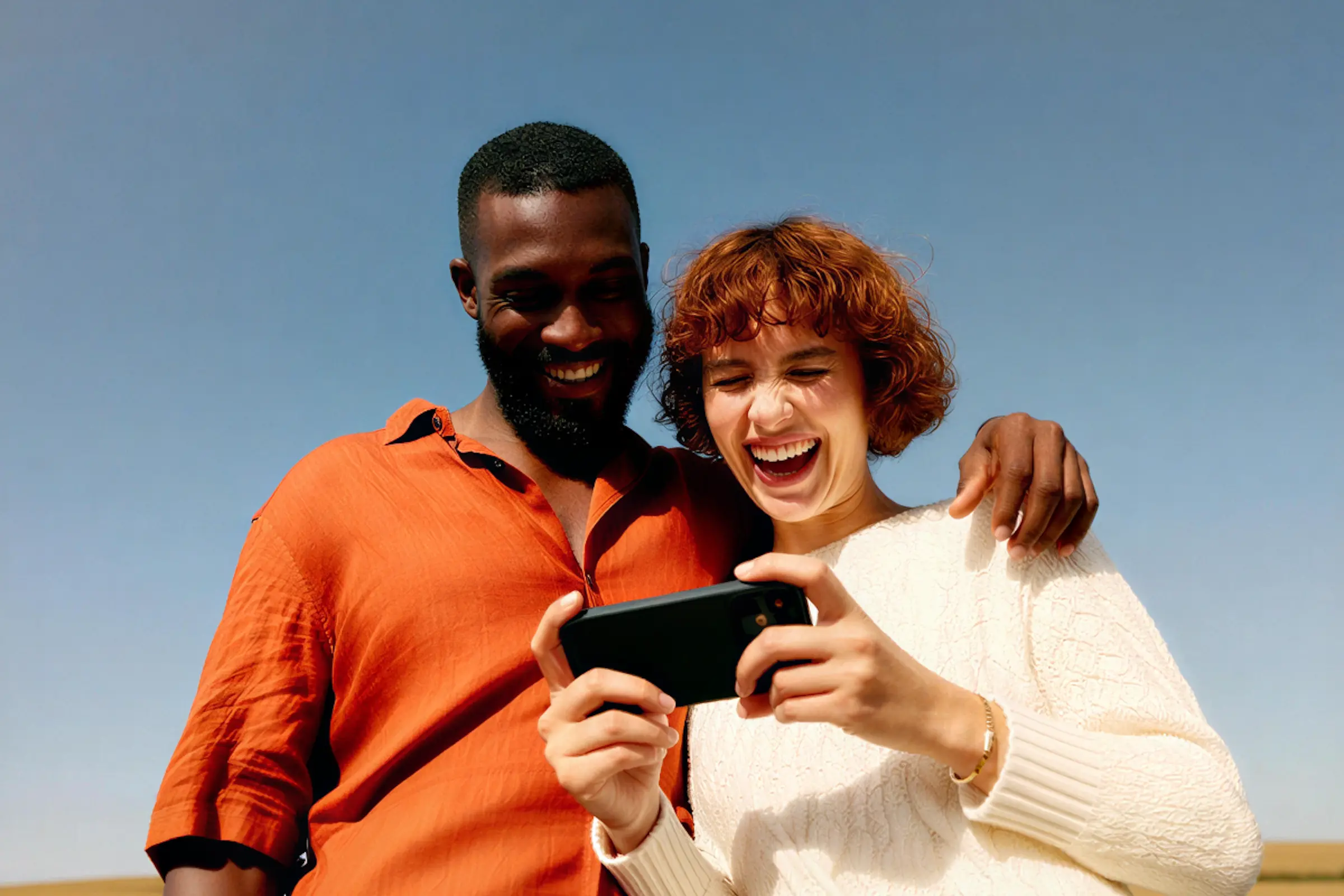 Smiling couple looking at a smartphone together outdoors under a clear sky.