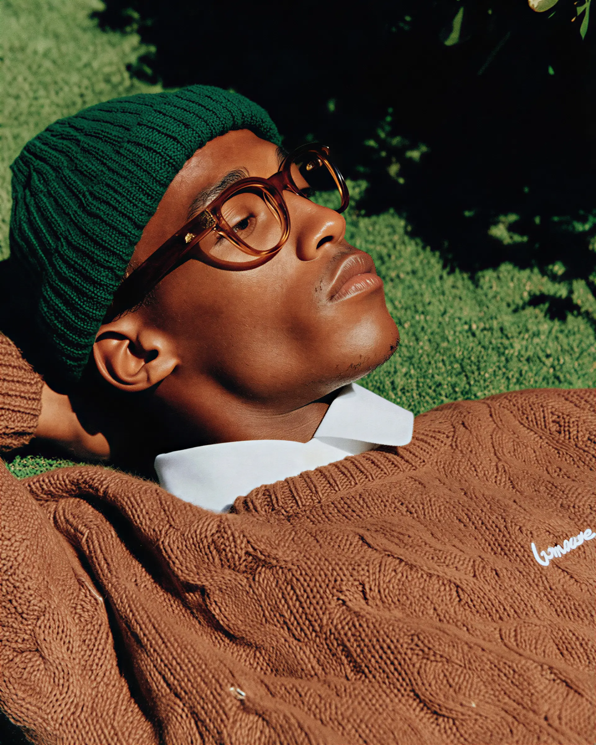 Man wearing a green knit beanie and brown cable knit sweater relaxing on grass with eyes closed.