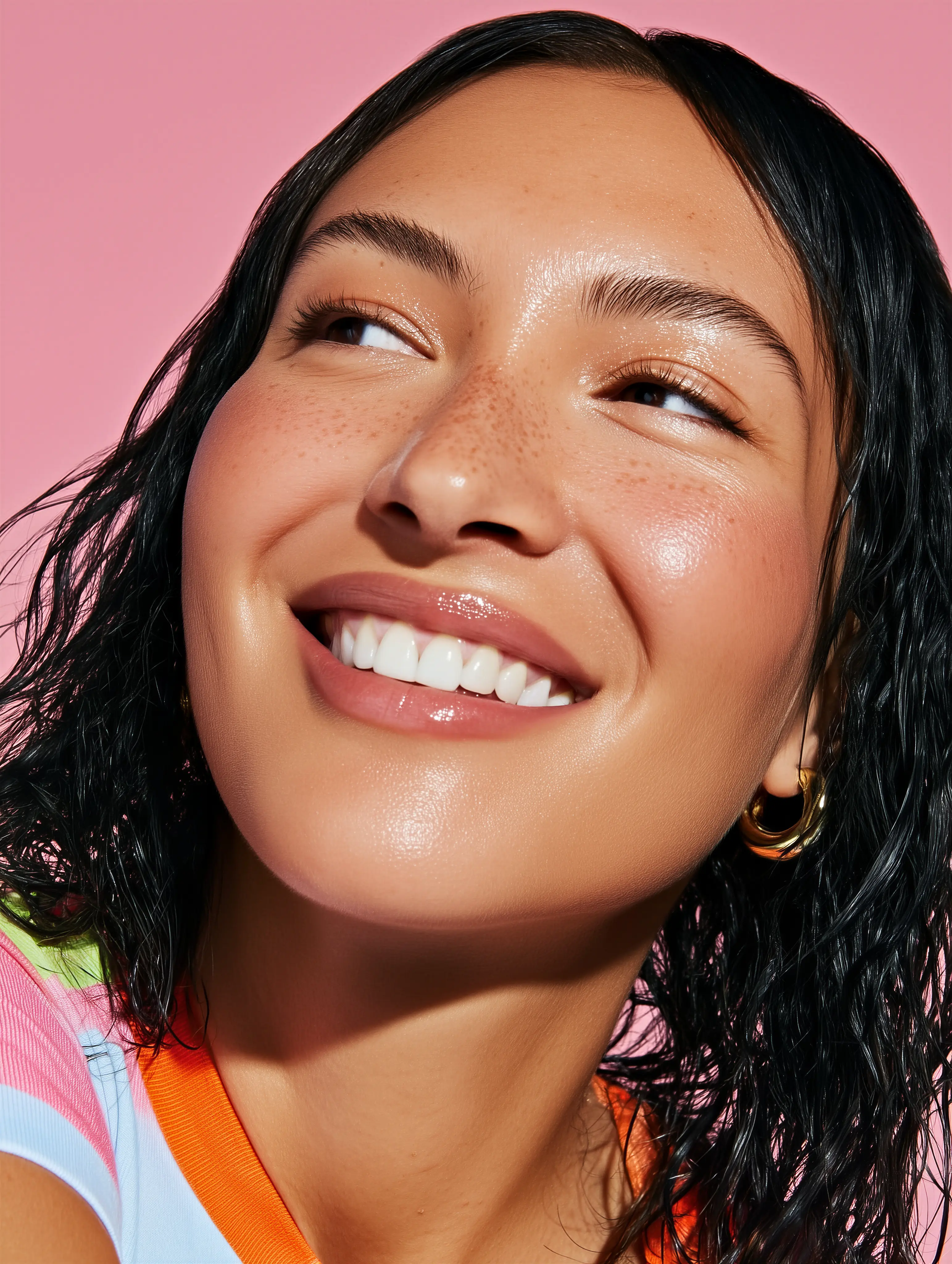 Close-up of a smiling woman with wet black hair, gold hoop earrings, and natural makeup against a pink background.