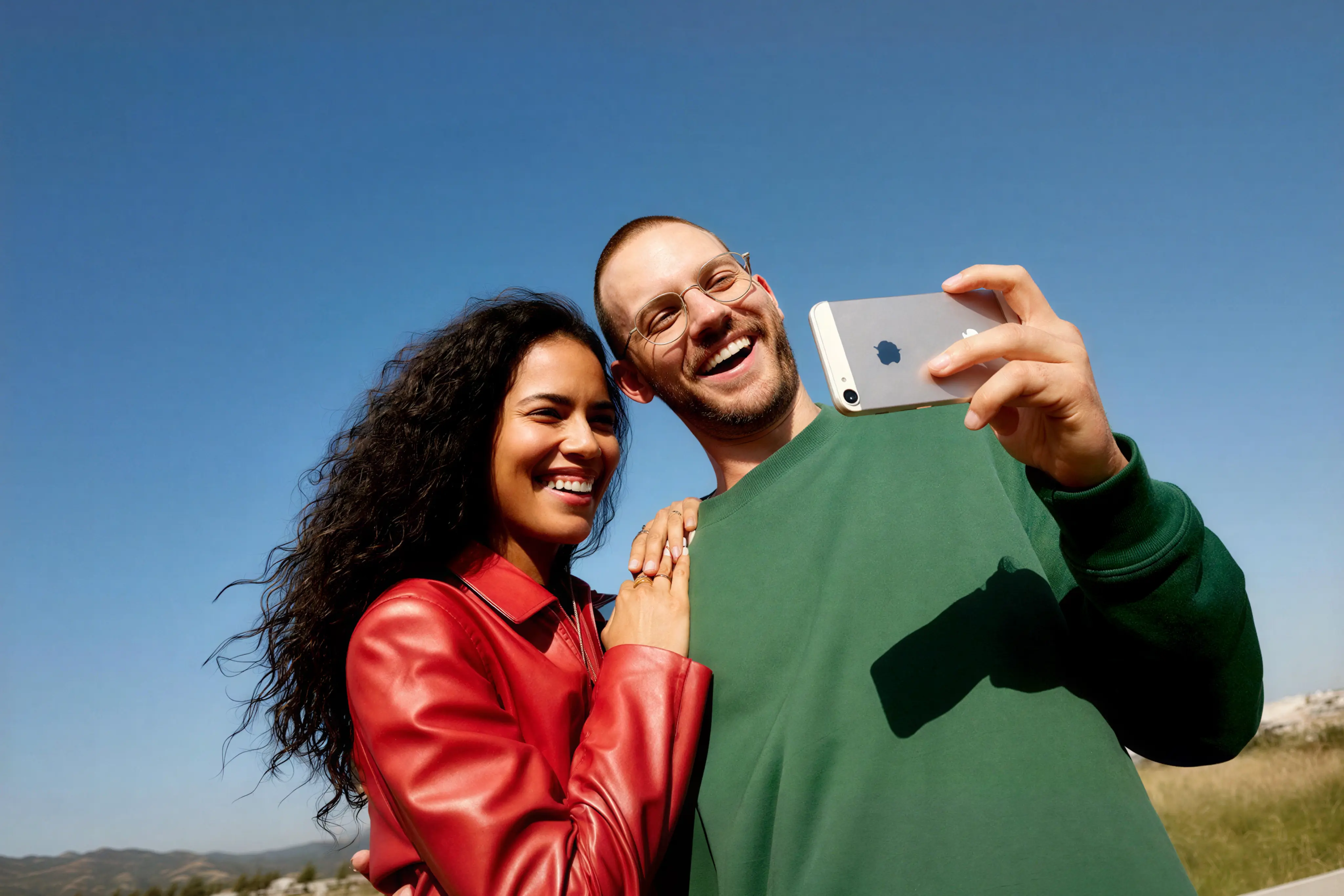 Smiling man in glasses and green sweater taking an outdoor selfie with woman in a red leather jacket against clear blue sky.