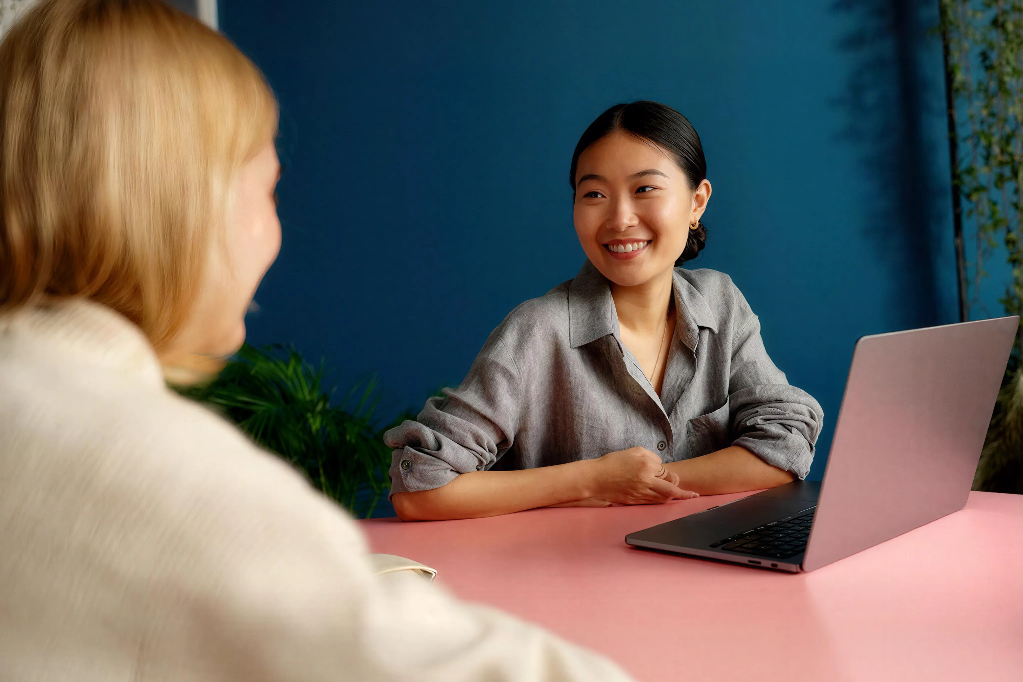 Two women having a friendly conversation at a pink table with a laptop, one smiling and listening attentively.