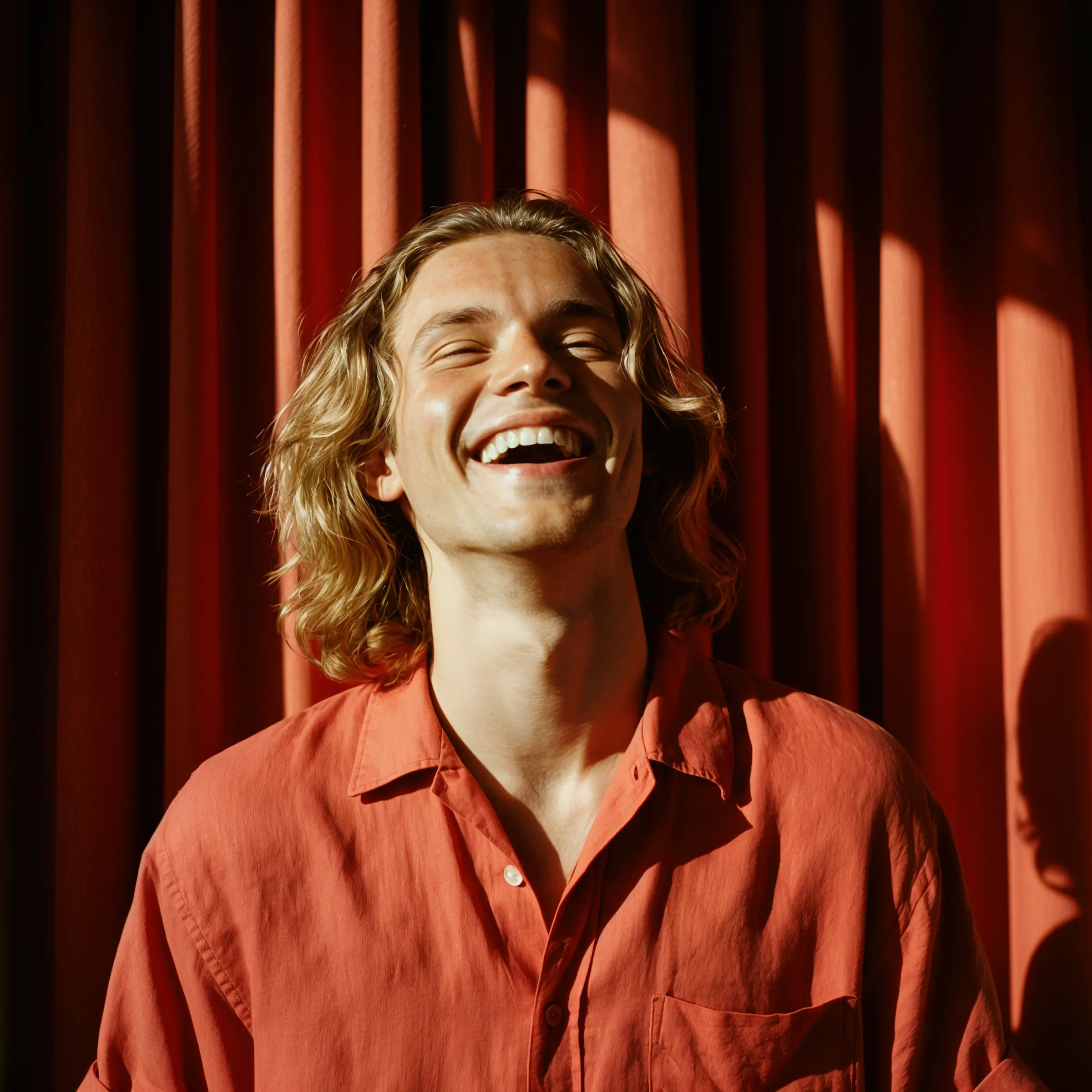 Smiling young man with blond hair in an orange shirt standing in front of red curtains.