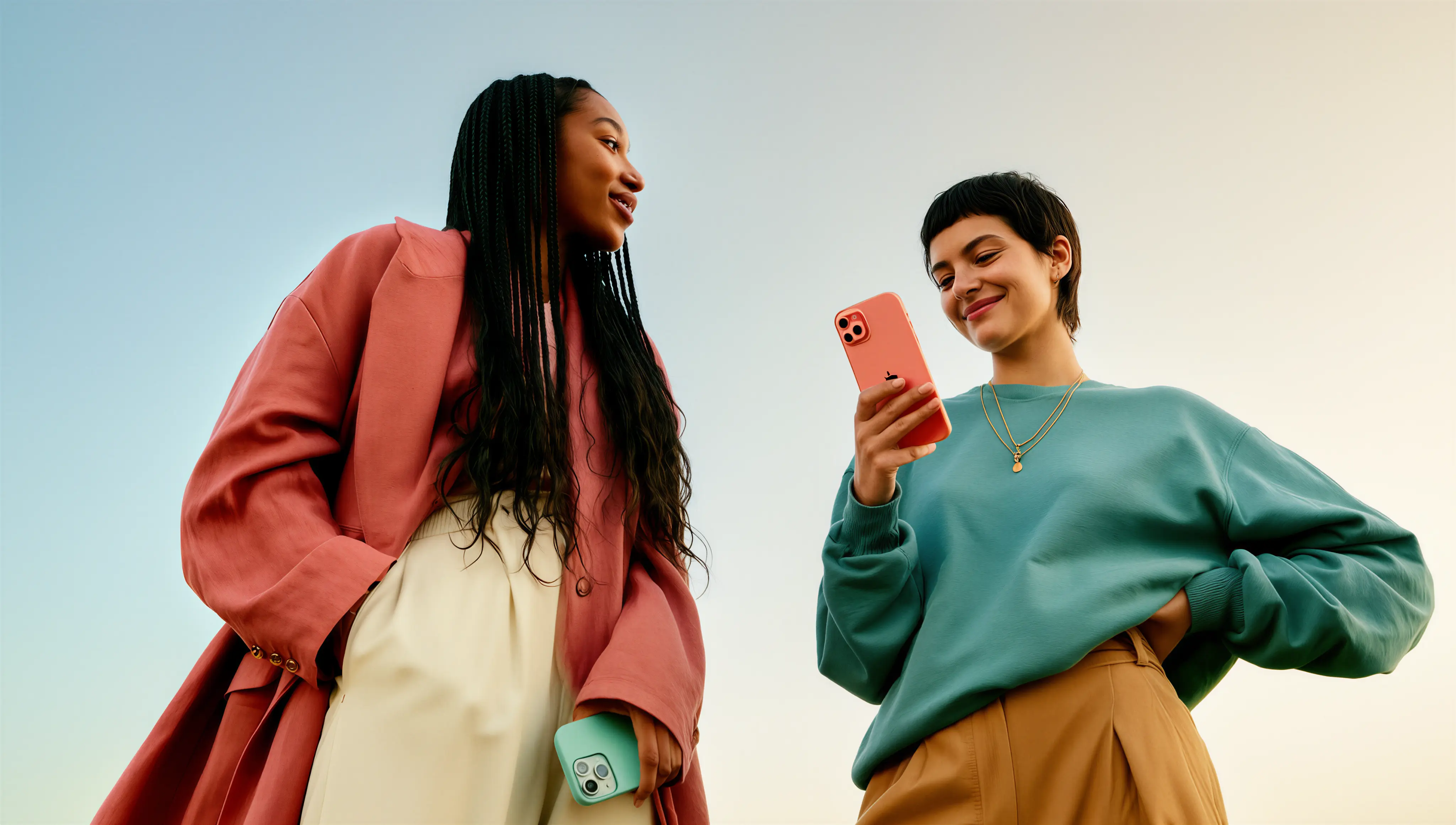 Two young women outdoors, one looking at her phone smiling while the other holds a phone and looks at her.