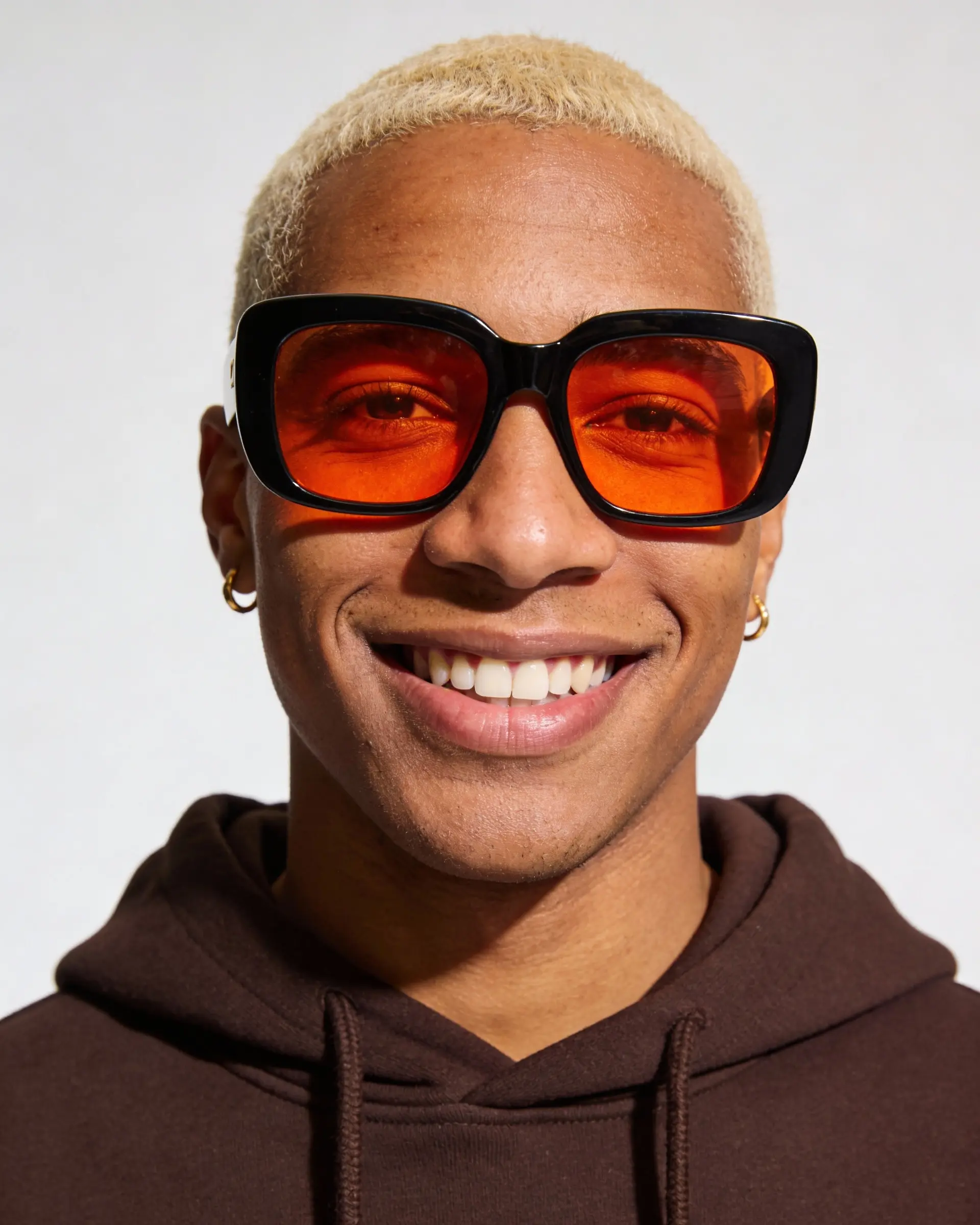 Smiling young person with short blond hair wearing large black-framed orange sunglasses and a dark hoodie.