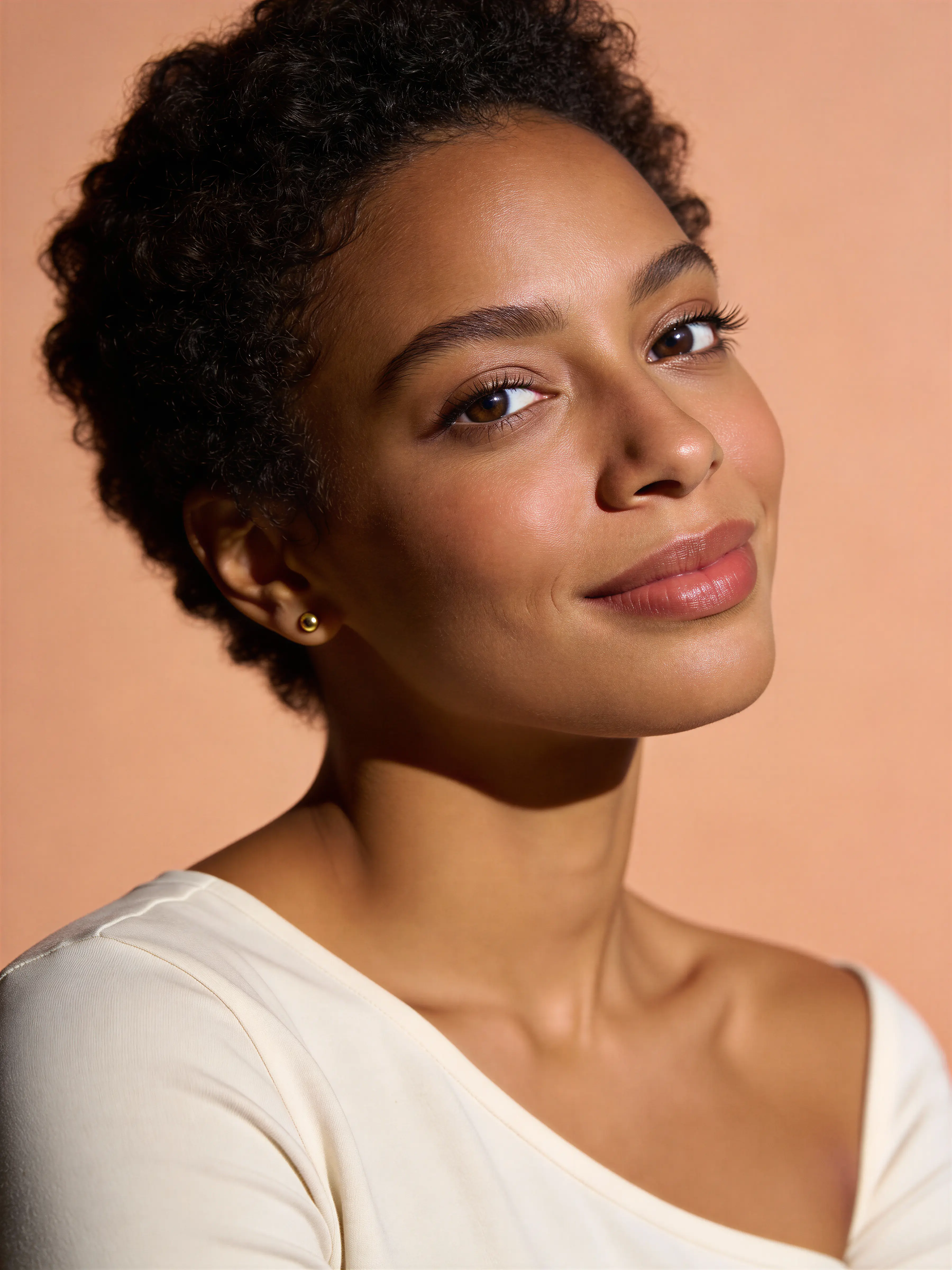 Portrait of a smiling woman with short curly hair wearing a cream-colored top against a peach background.