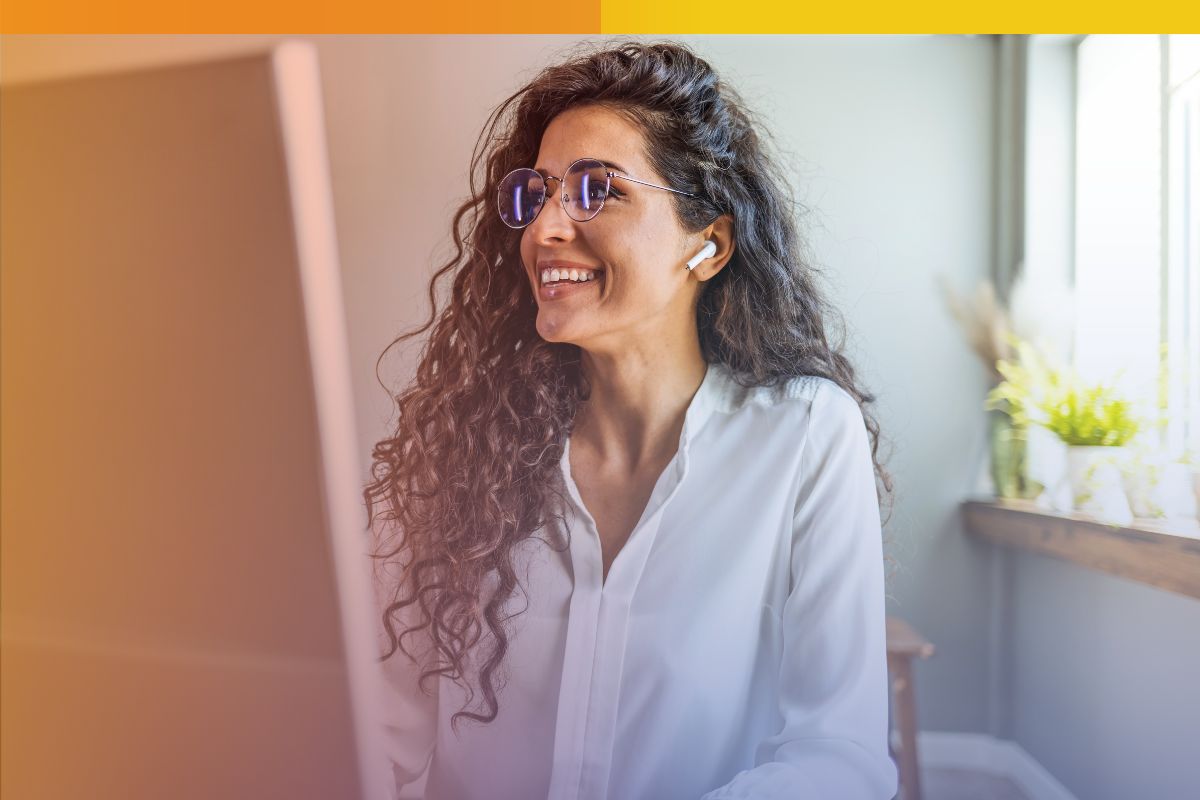 A smiling content moderator with curly hair and glasses looking at a computer screen with wireless earbuds, illustrating Connext Global Solutions
