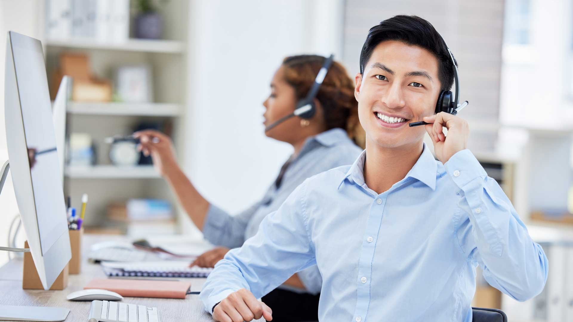 Smiling customer support professional wearing a headset at a computer workstation, with another headset-wearing colleague working in the background.|How to Outsource IT Support Without Losing Control of Service Quality