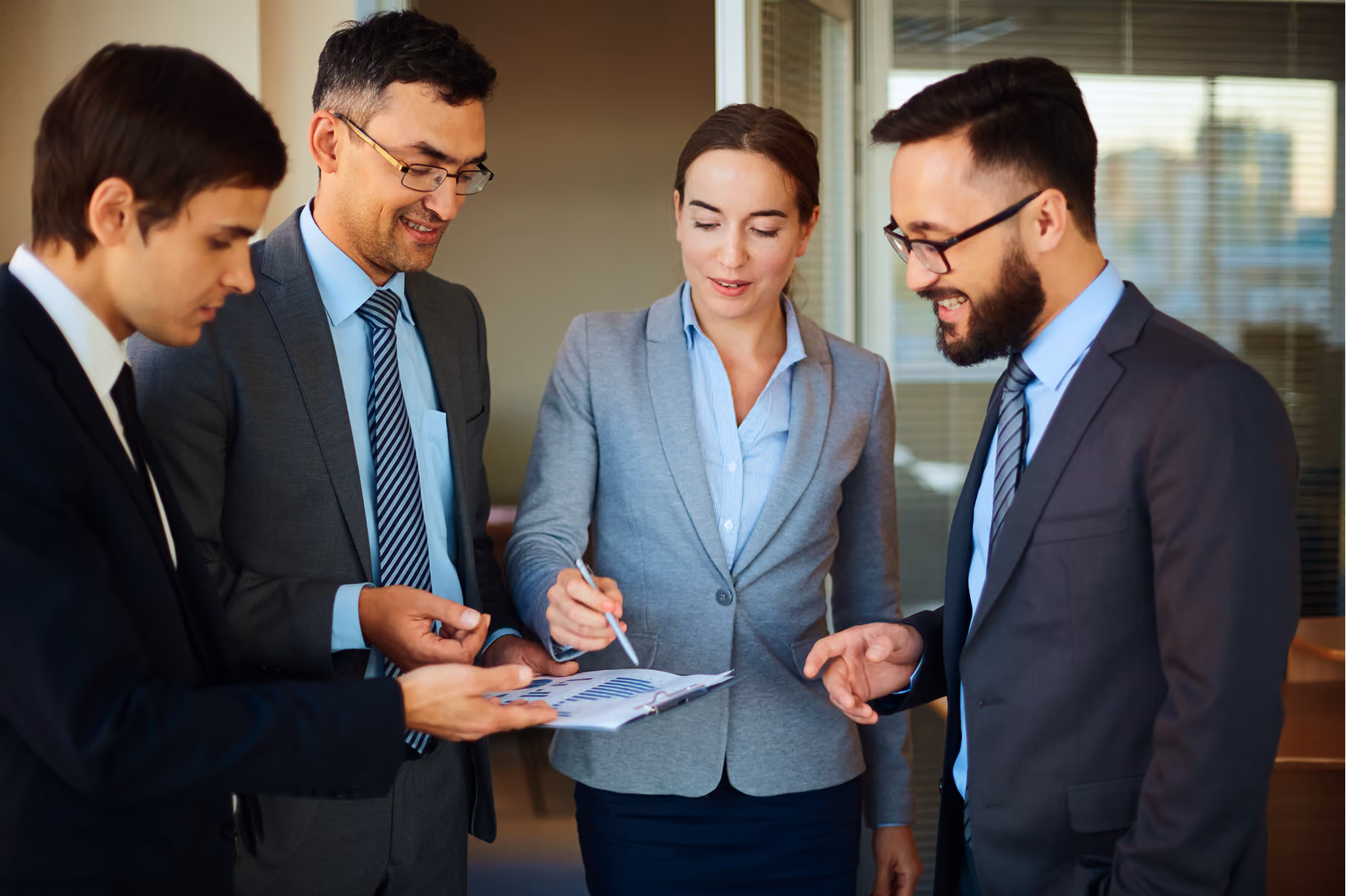 Quatre professionnels en costume debout dans un bureau, examinant et pointant des graphiques de données sur un presse-papiers.